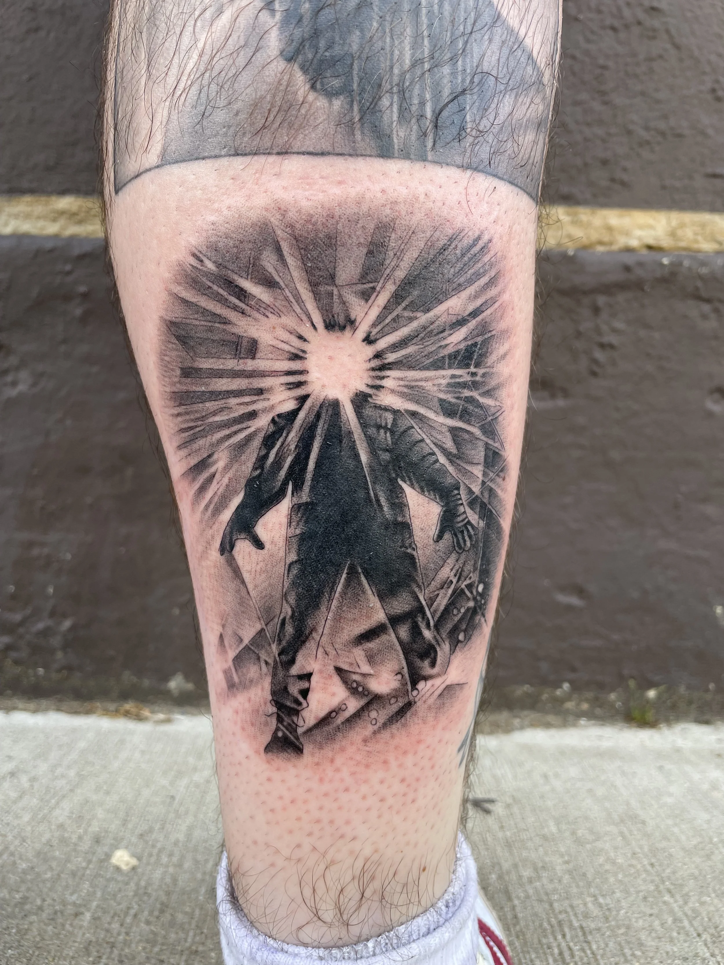 Tattoo of a man with a shattered bright light behind him, standing with arms outstretched and sheets of glass breaking around him, on a person's leg.