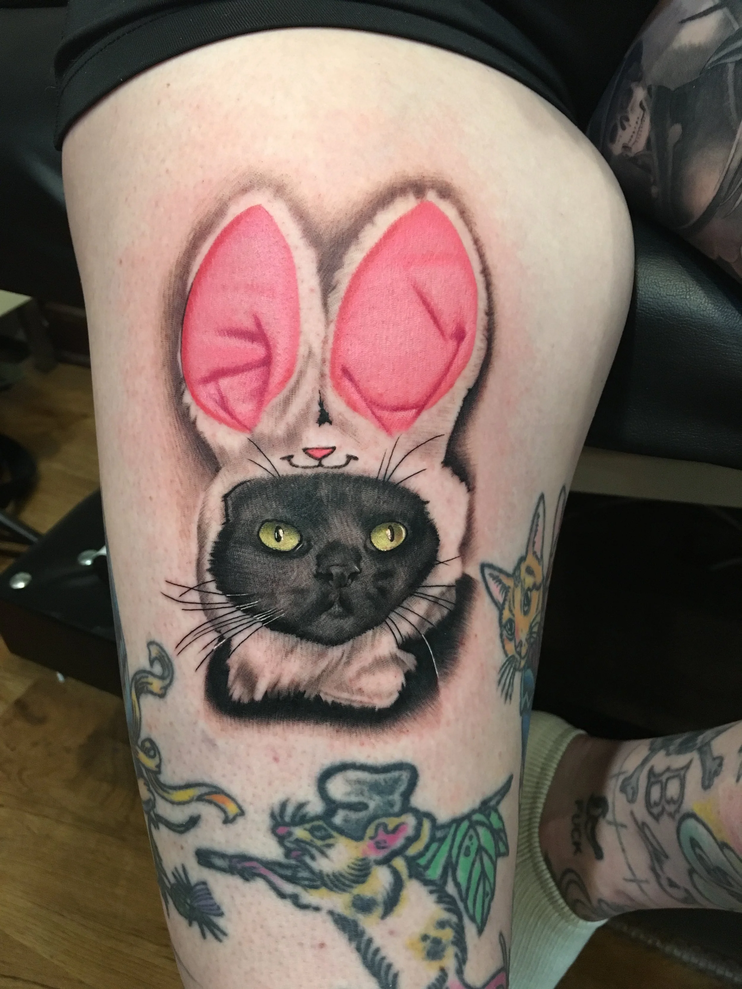 Tattoo of a black cat's face with yellow eyes, with a bunny outline with pink ears and closed eyes above it, on person's thigh.