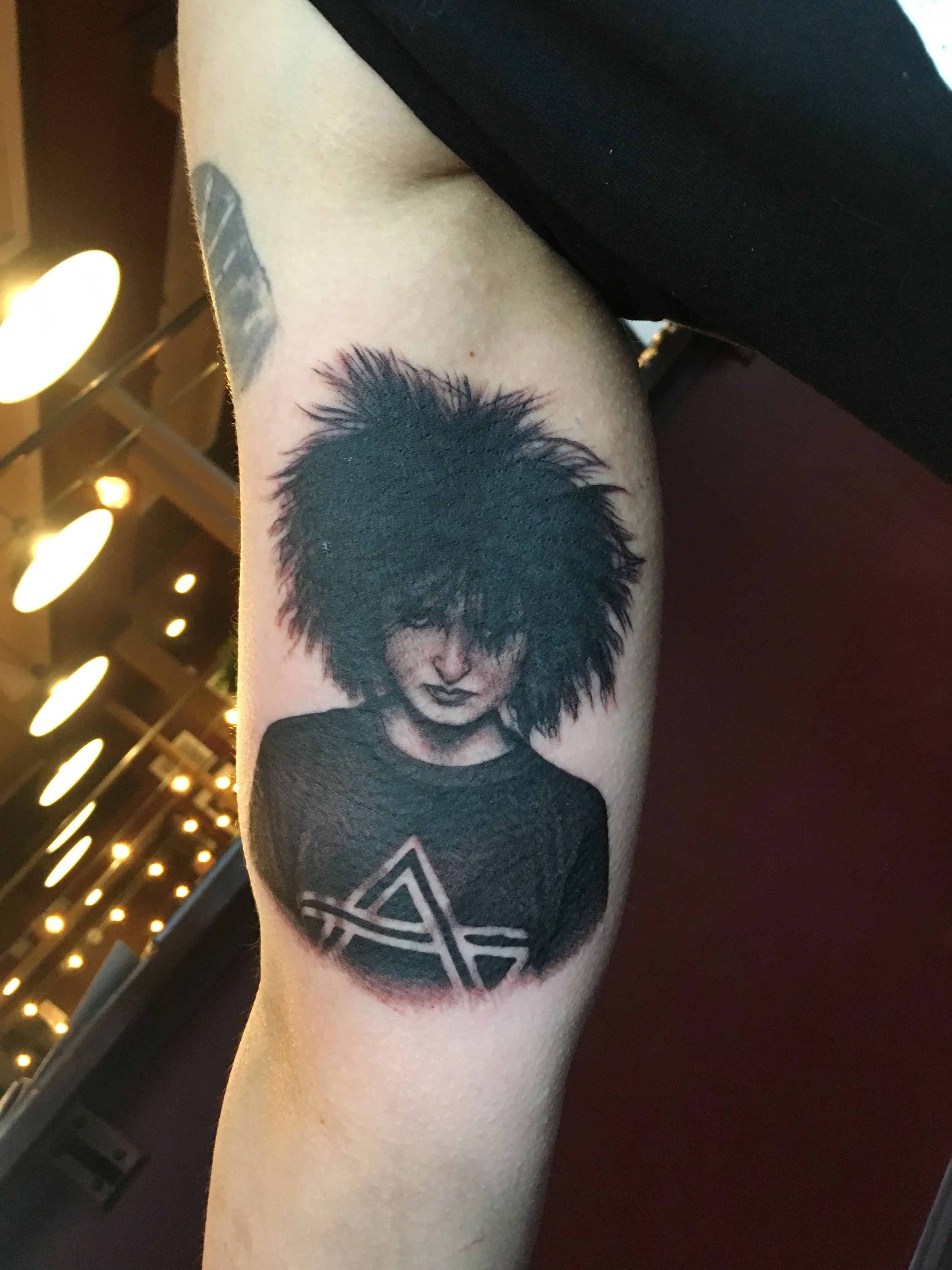 Tattoo of a person with messy black hair, wearing a dark shirt with a geometric design, on person's upper arm.
