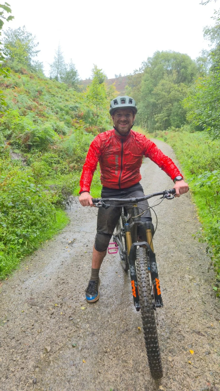 A man in a red windbreaker and black shorts riding a mountain bike on a dirt trail in a lush green forest, wearing a helmet and smiling.