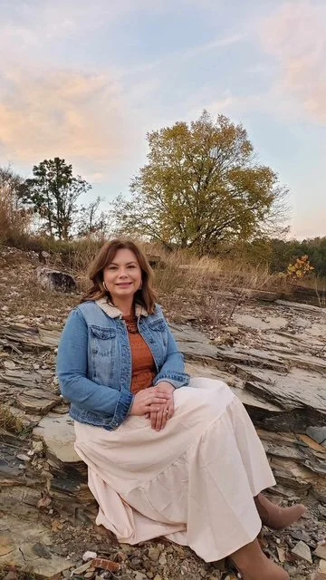 A woman sitting outdoors on rocky terrain near a large tree, wearing a denim jacket and a long skirt, smiling at the camera during sunset or late afternoon.