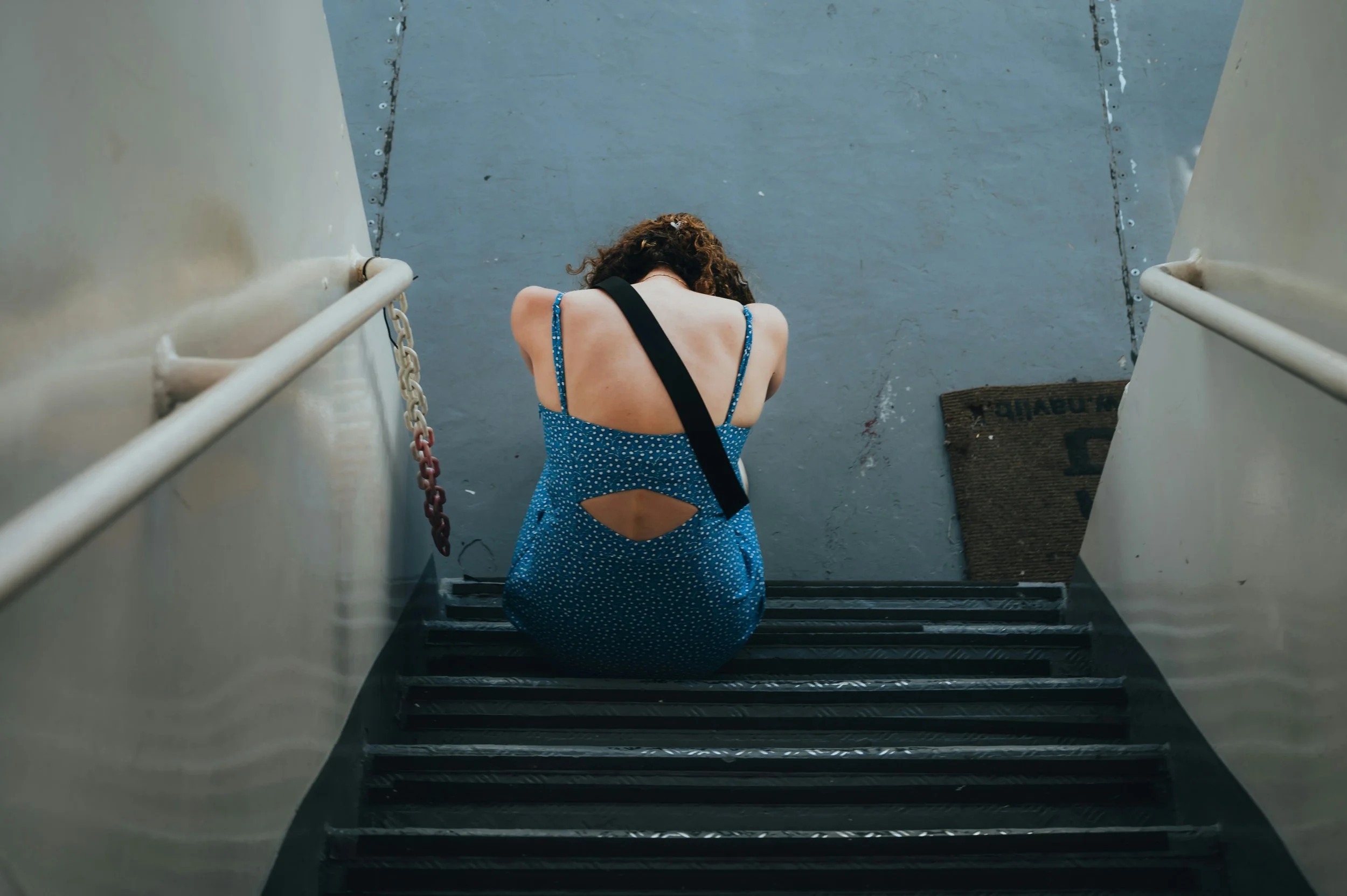 A woman in a blue dress with white polka dots sitting on stairs, with her head bowed and arms crossed over her knees, at the bottom of a staircase.