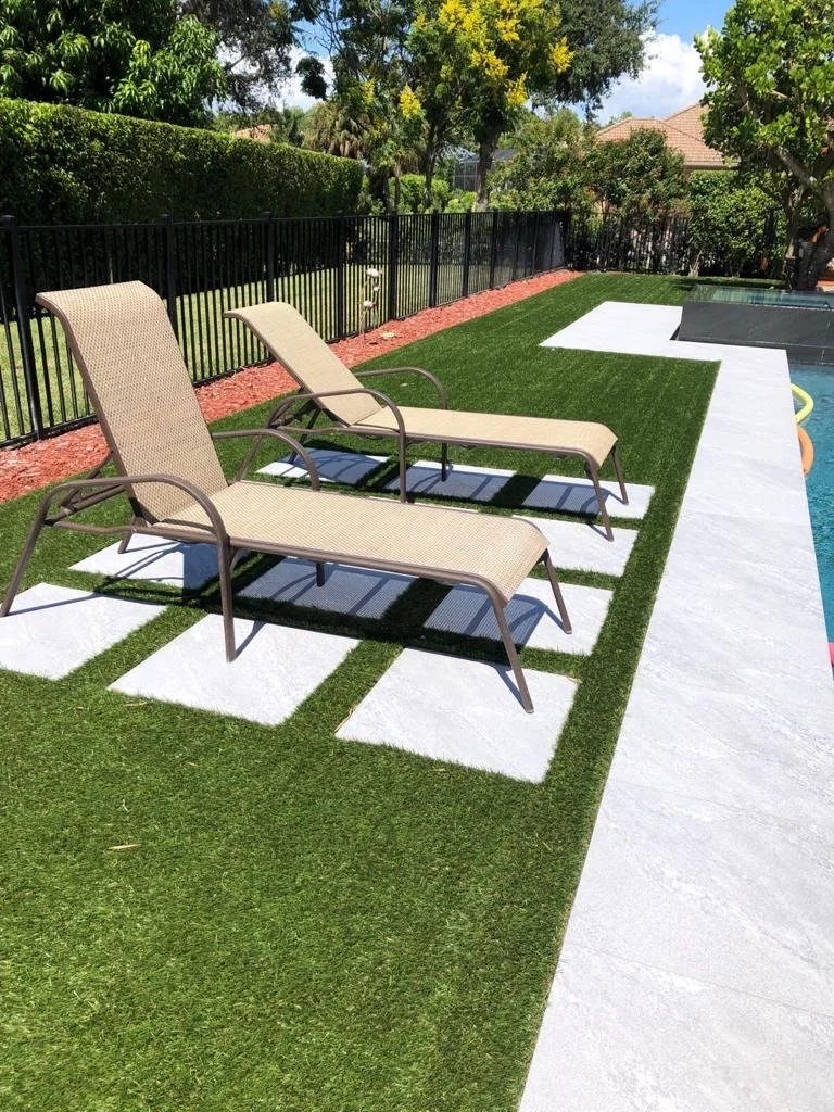 Two beige patio lounge chairs on a well-manicured lawn beside a swimming pool, enclosed by a black metal fence, with trees and shrubbery in the background.