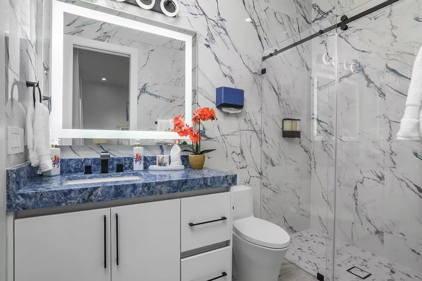 Modern bathroom with marble walls and a blue marble counter, white vanity and mirror, toilet, floral decorations, and a shower area with a glass partition.