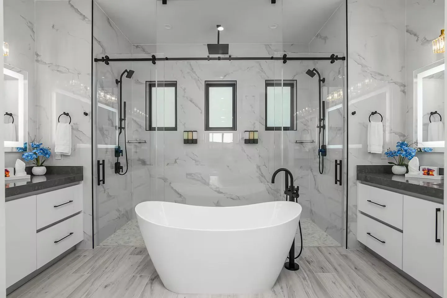 Modern bathroom with a freestanding bathtub in the center, surrounded by white marble walls and floors, two black showerheads, and dual vanities with mirrors, flowers, and toiletries.