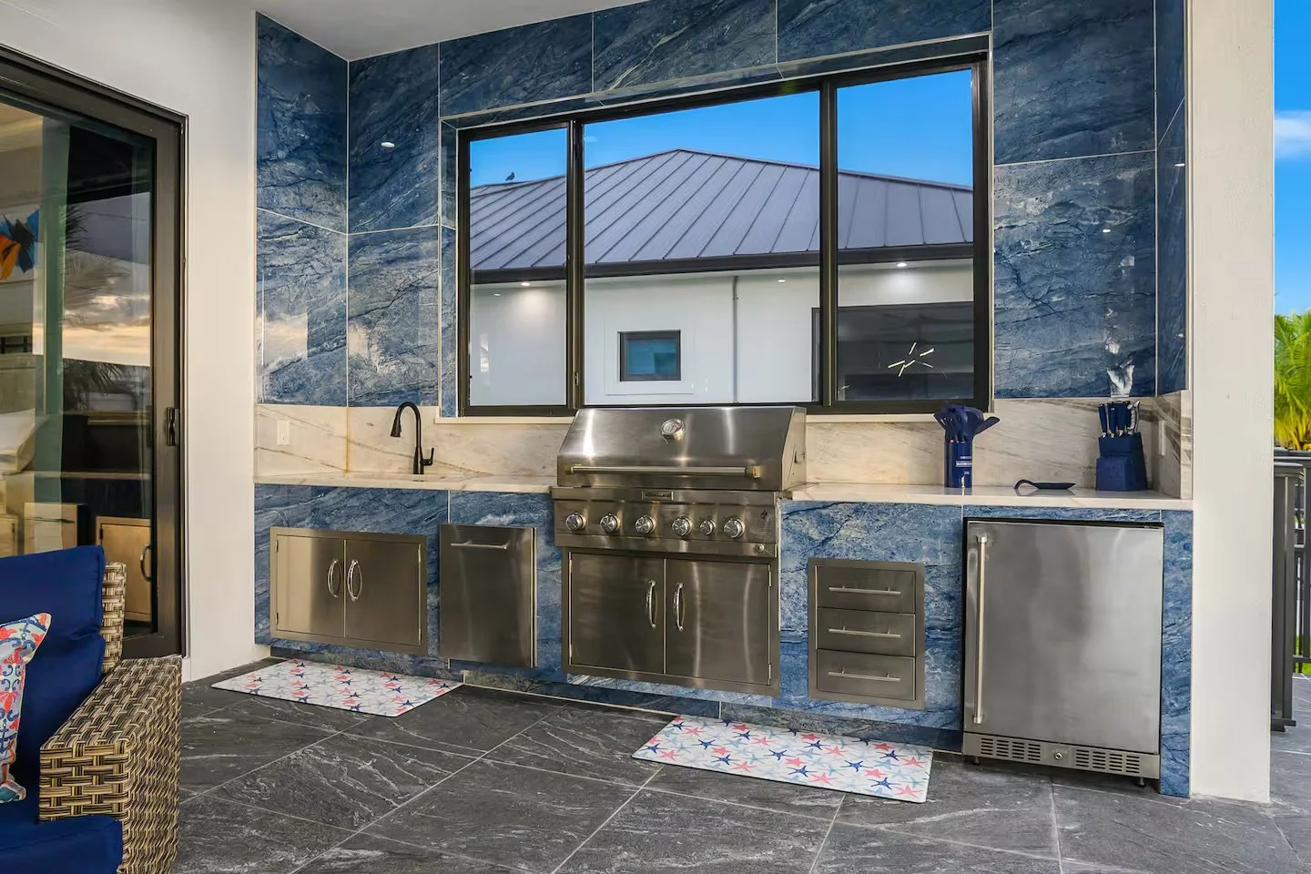 Outdoor kitchen with a stainless steel grill, sink, and small fridge, surrounded by blue and white marble tiles, on a patio with dark gray stone flooring.