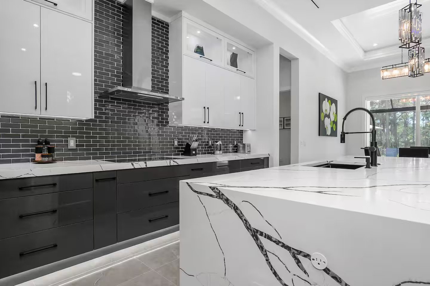 Modern kitchen with black and white cabinetry, black subway tile backsplash, white marble countertops, and a large white island with black veining. Stainless steel appliances, black fixtures, and decorative lighting fixtures hang from the ceiling. La