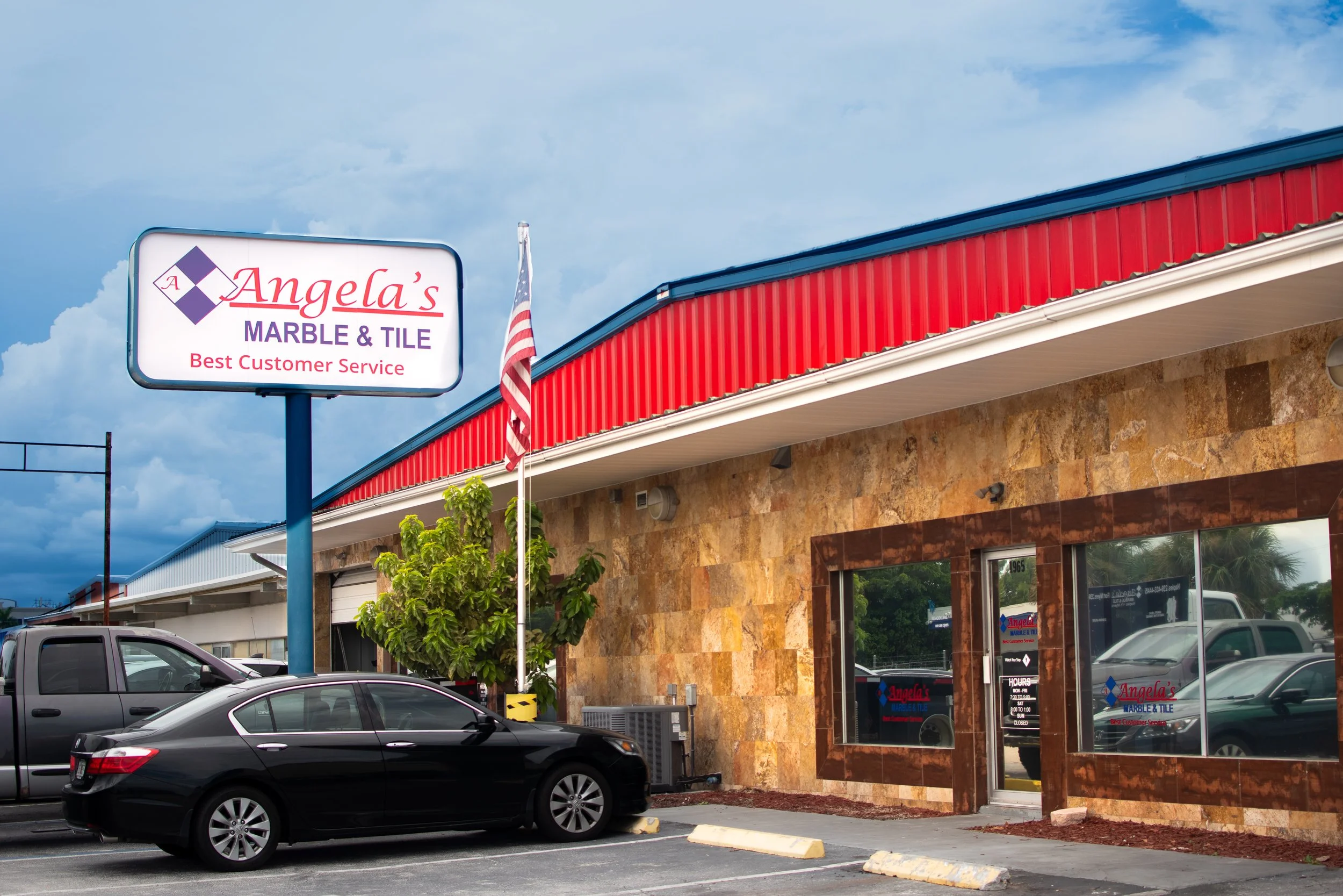 Front view of Angela's Marble & Tile store with a parking lot, a black car parked in front, a large sign, American flag, and a red and blue building exterior.