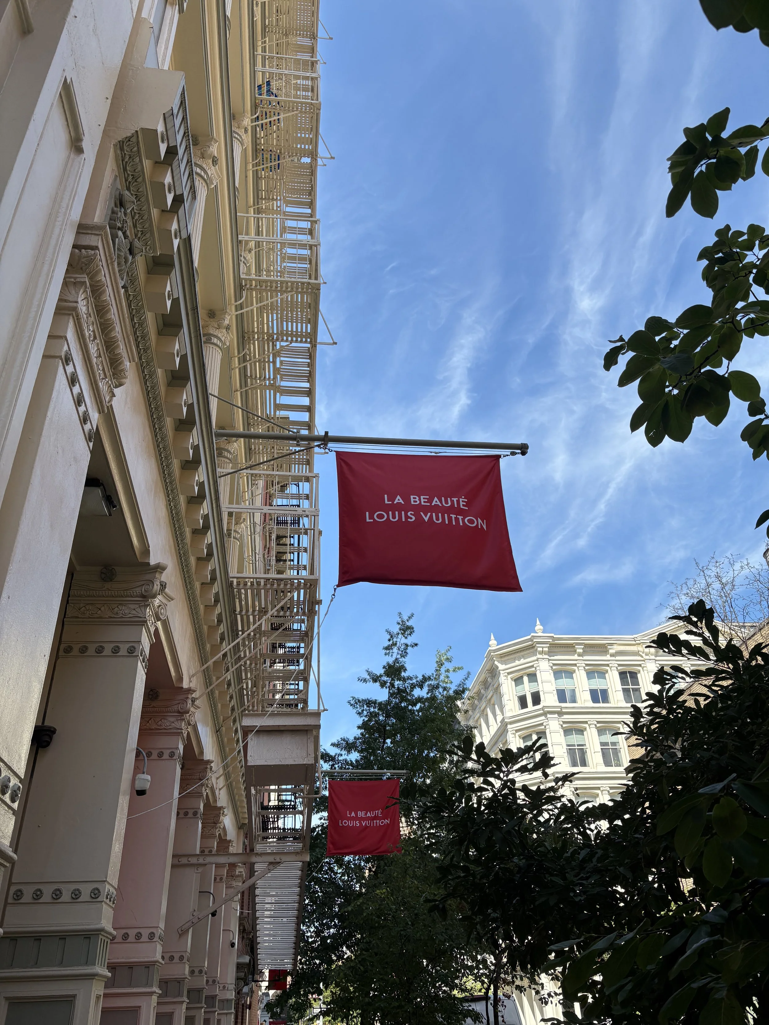 Two red flags with white text reading 'LA BEAUTÉ LOUIS VUITTON' hang outside a beige building with ornate architectural details, trees, and a blue sky.