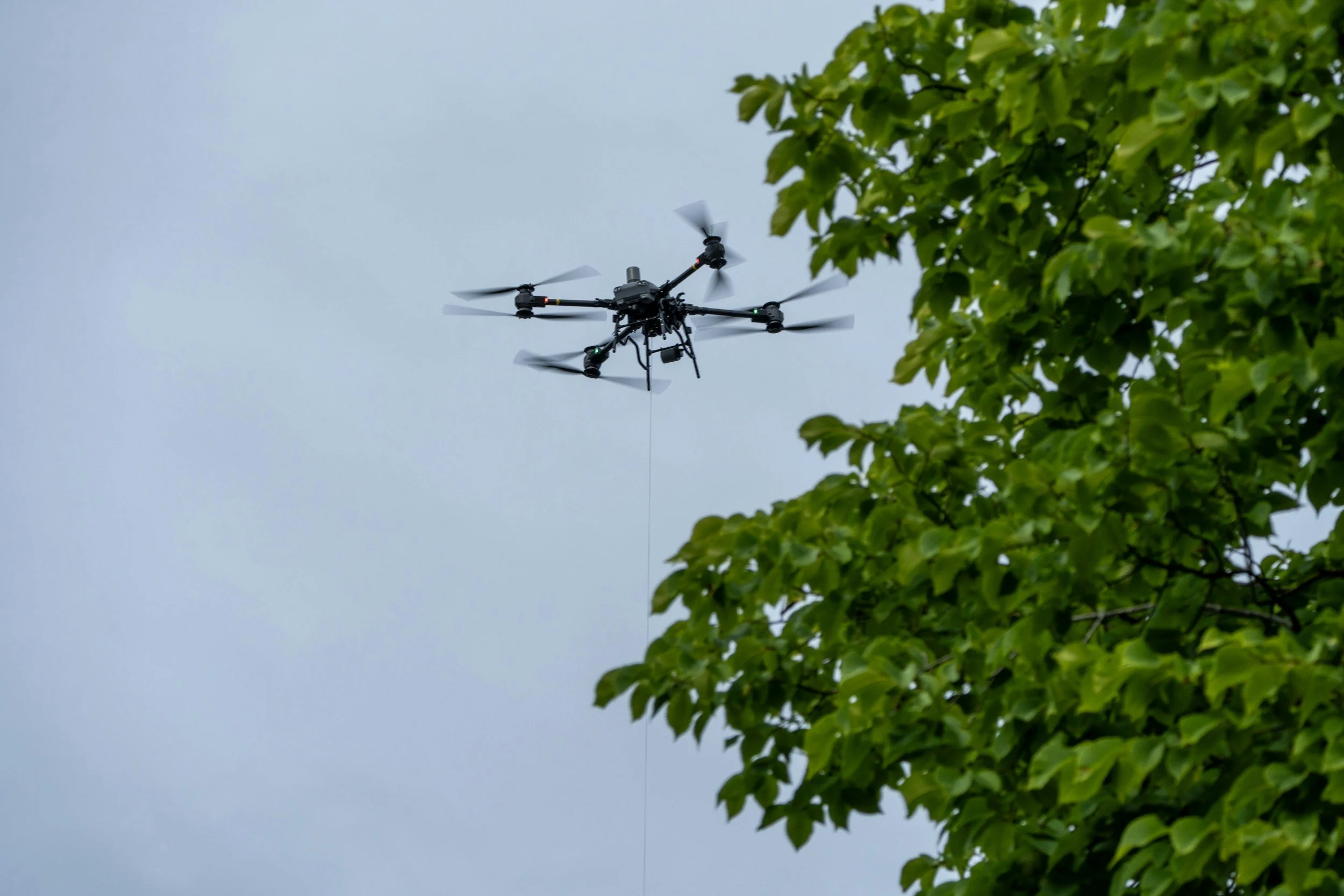 A lifting drone flying in the sky next to green tree leaves.