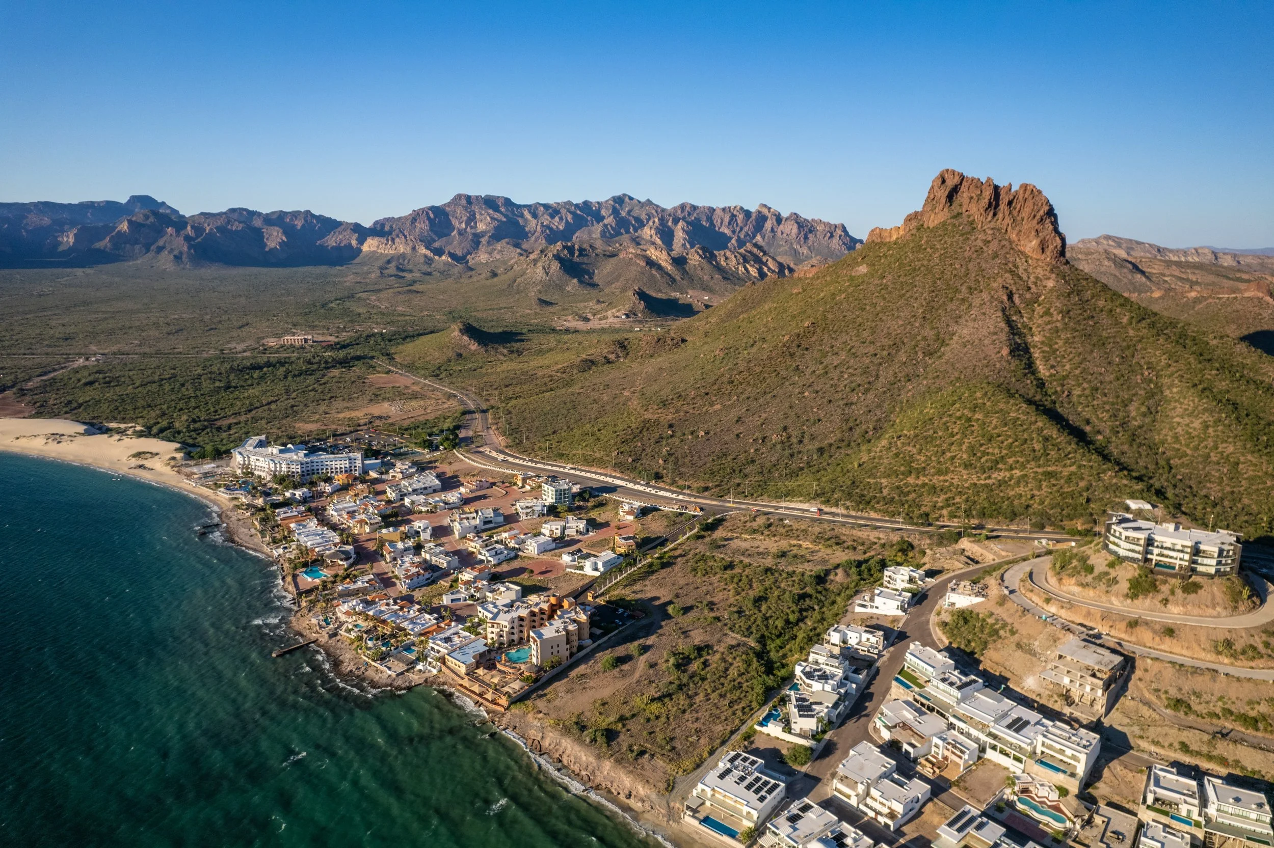 Vista aérea de un pueblo junto a la costa con montañas en el fondo, incluyendo una formación rocosa prominente.