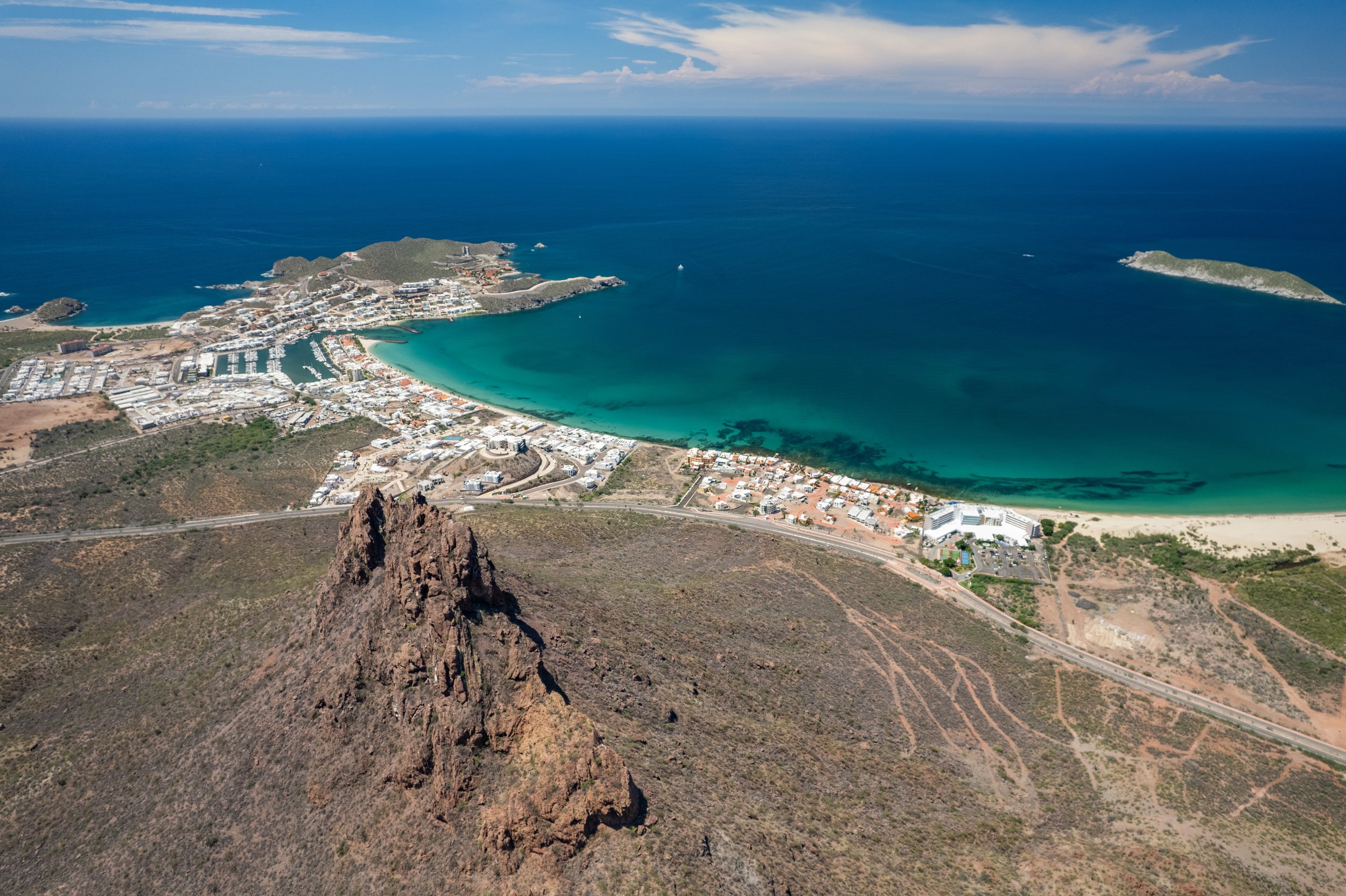 Vista aérea de una playa en forma de medialuna con aguas turquesa, rodeada de una zona urbana y un paisaje montañoso con rocas y vegetación seca.