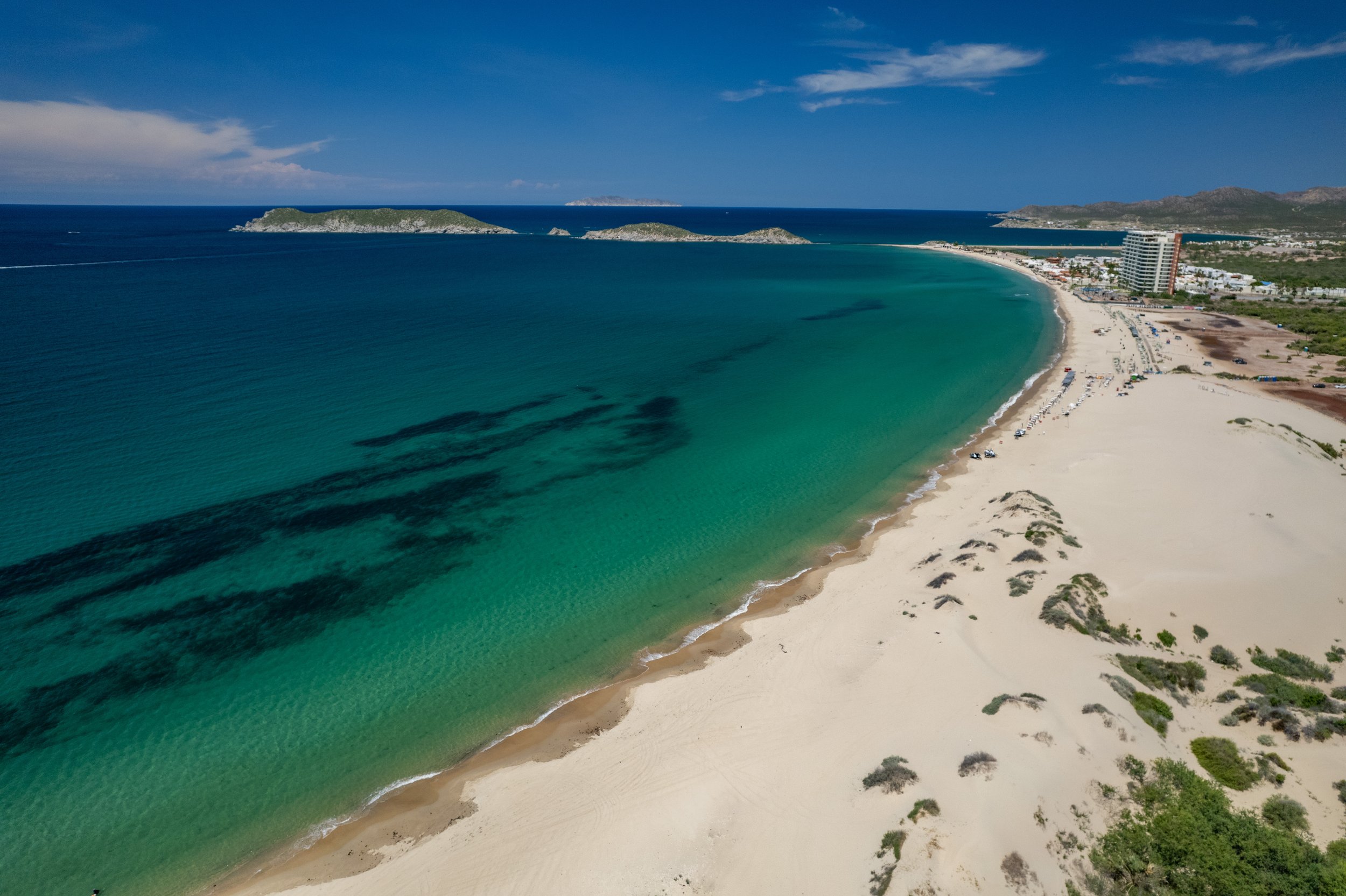 Vista panorámica de una playa con arena clara y agua turquesa, con algunas zonas sombreadas en el mar, y edificios y colinas en el fondo.