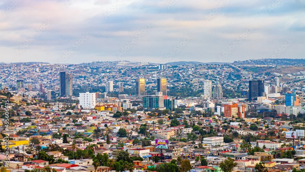 Vista panorámica de un paisaje urbano con edificios altos y casas pequeñas, con colinas al fondo bajo un cielo nublado.
