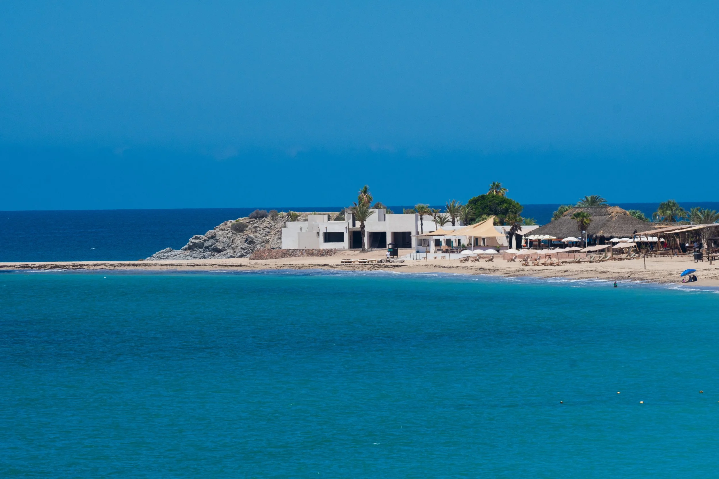 Vista de una playa con agua azul clara, arena y estructuras de playa con palapas y algunas plantas de palmera, al fondo se observa un edificio blanco y una roca