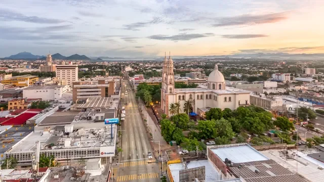 Vista aérea de un centro urbano con un edificio emblemático con cúpula y campanario en el centro, rodeado de árboles, con calles y otros edificios a su alrededor durante el atardecer, y montañas en el fondo.