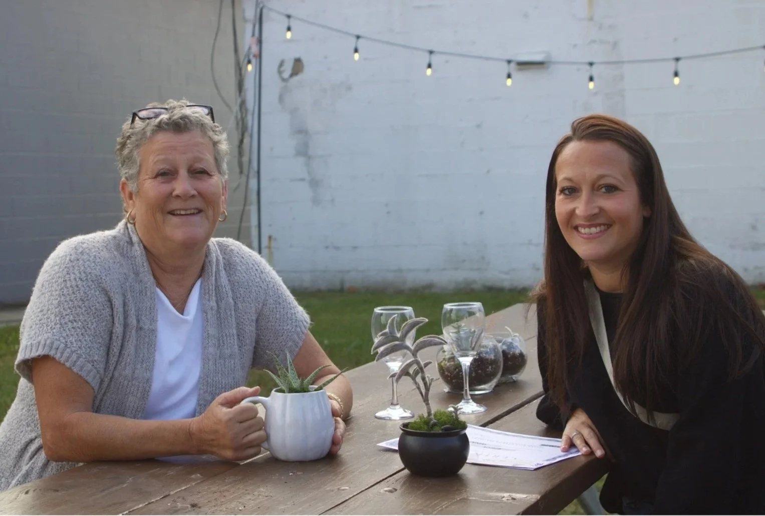 Two women sitting at a wooden table outdoors in a backyard, smiling. The older woman on the left holds a white mug with a plant, and the younger woman on the right has a paper in front of her. There are three wine glasses with drinks and small potted plants on the table, with string lights hanging above in the background.