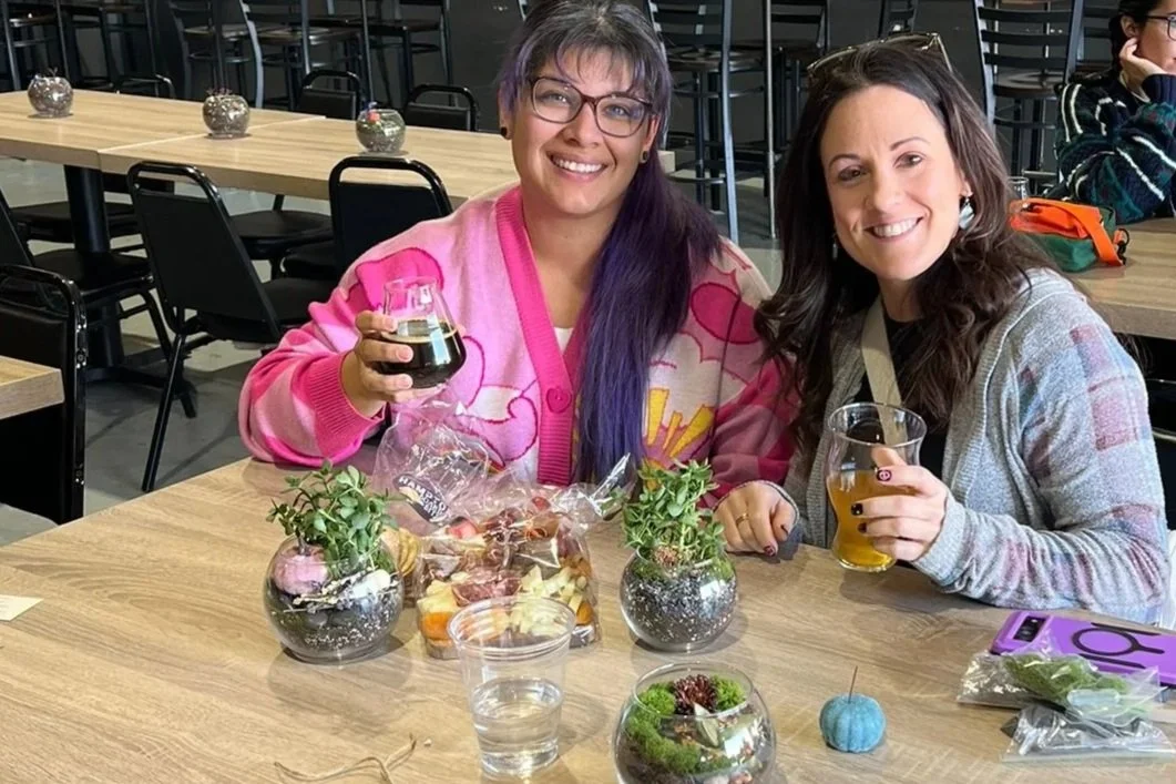 Two women sitting at a table, smiling and holding drinks, with small potted plants and a bag of snacks on the table, in a room with empty chairs and tables in the background.