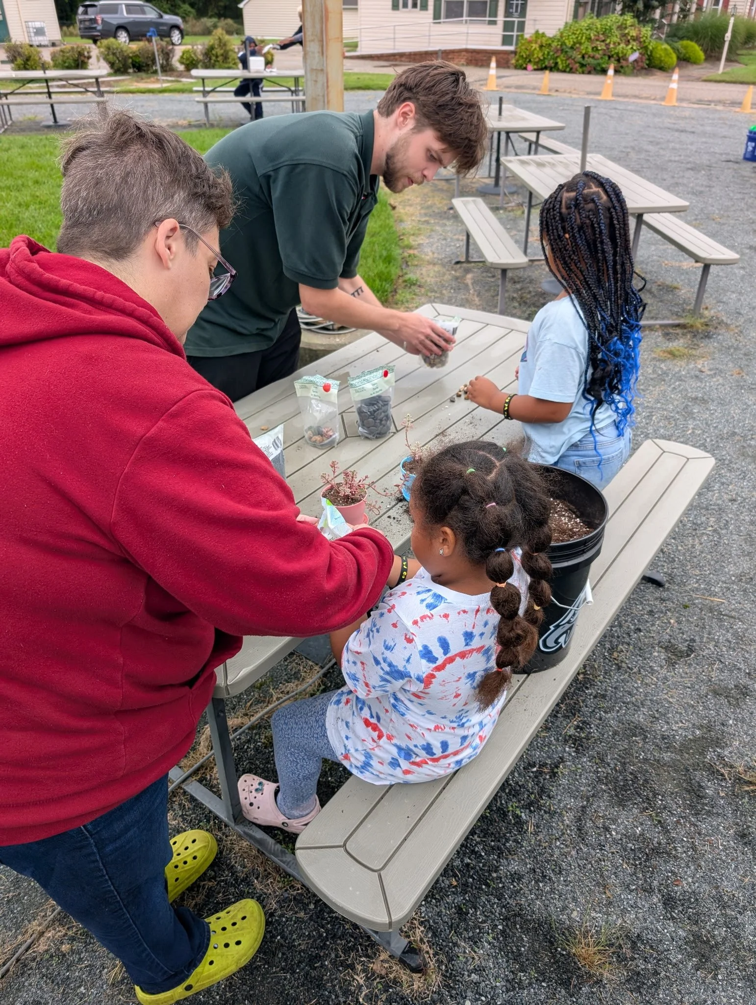 Two adults and two children planting flowers in pots outdoors at a picnic table.