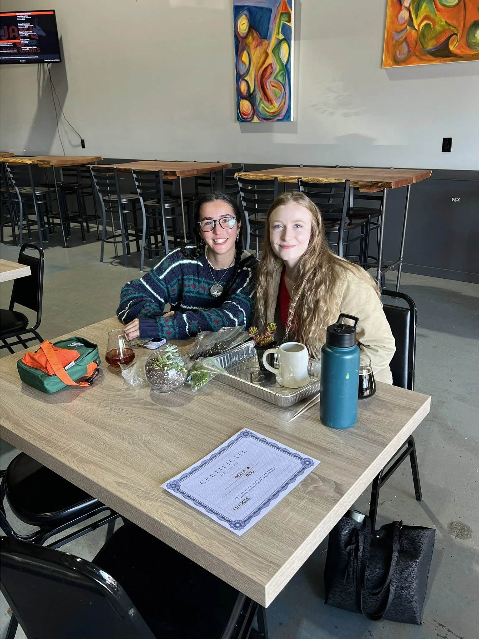 Two women sitting at a wooden table in a brightly lit restaurant or cafe, smiling at the camera. The table has a certificate, a water bottle, a coffee mug, a small plant, and some objects. The background features colorful abstract paintings on the wall and empty high-top tables with bar stools.