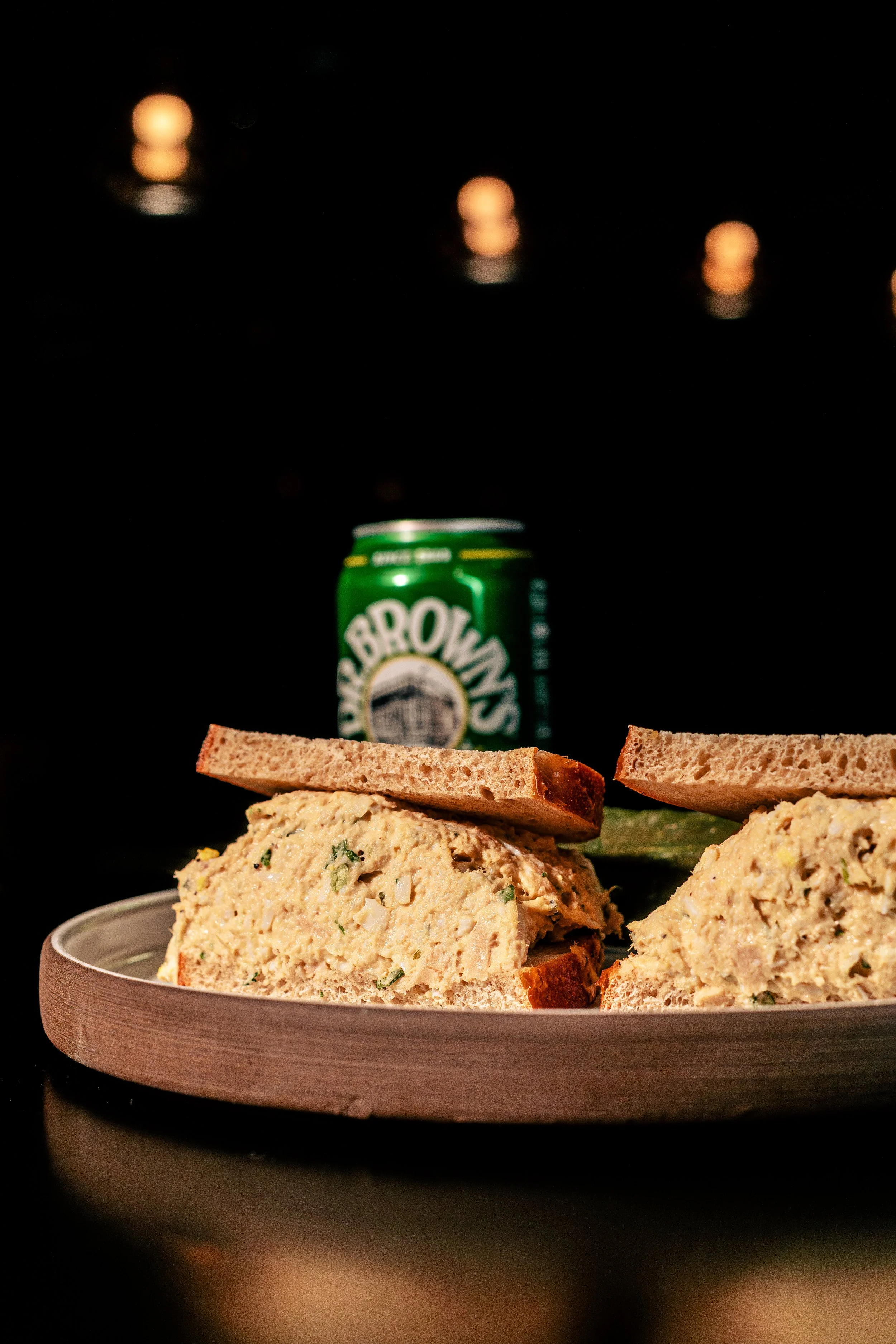 A plate with egg salad sandwiches and a can of Seven-Up soda in the background against a black backdrop with string lights.