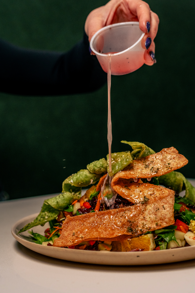 A person pouring sauce over a plate of fish tacos on a bed of vegetables with green peppers and tomatoes.