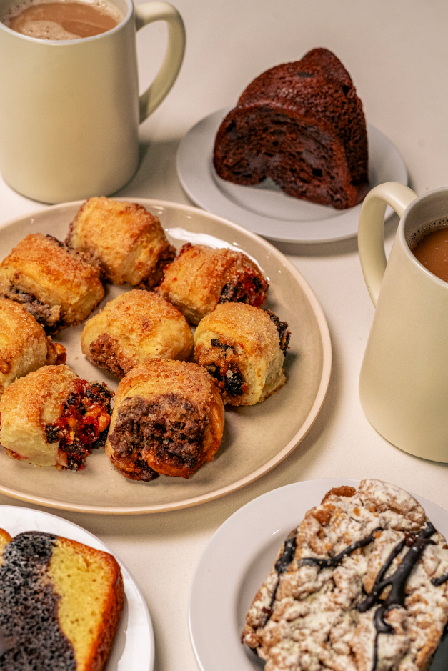 Plate of baked apple desserts with a filling of dark fruit, possibly berries, surrounded by a golden crust. Two mugs of hot chocolate or coffee, and slices of chocolate and fruit cakes. A plate with two slices of dark chocolate bread or cake in the b