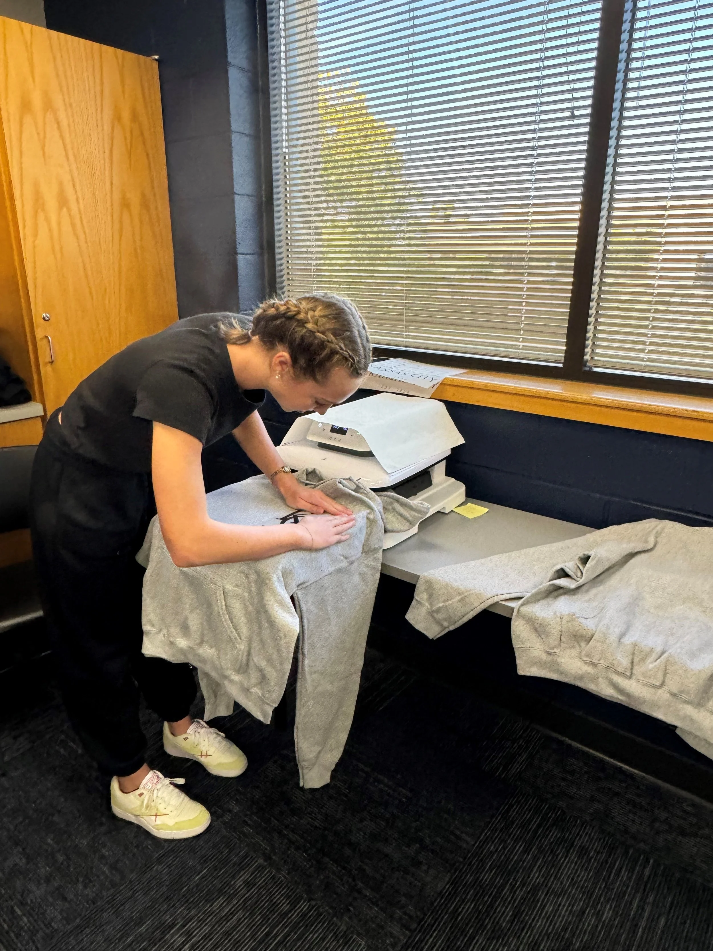 A woman ironing a gray sweatshirt on an ironing board in an office, with blinds covering a window in the background.