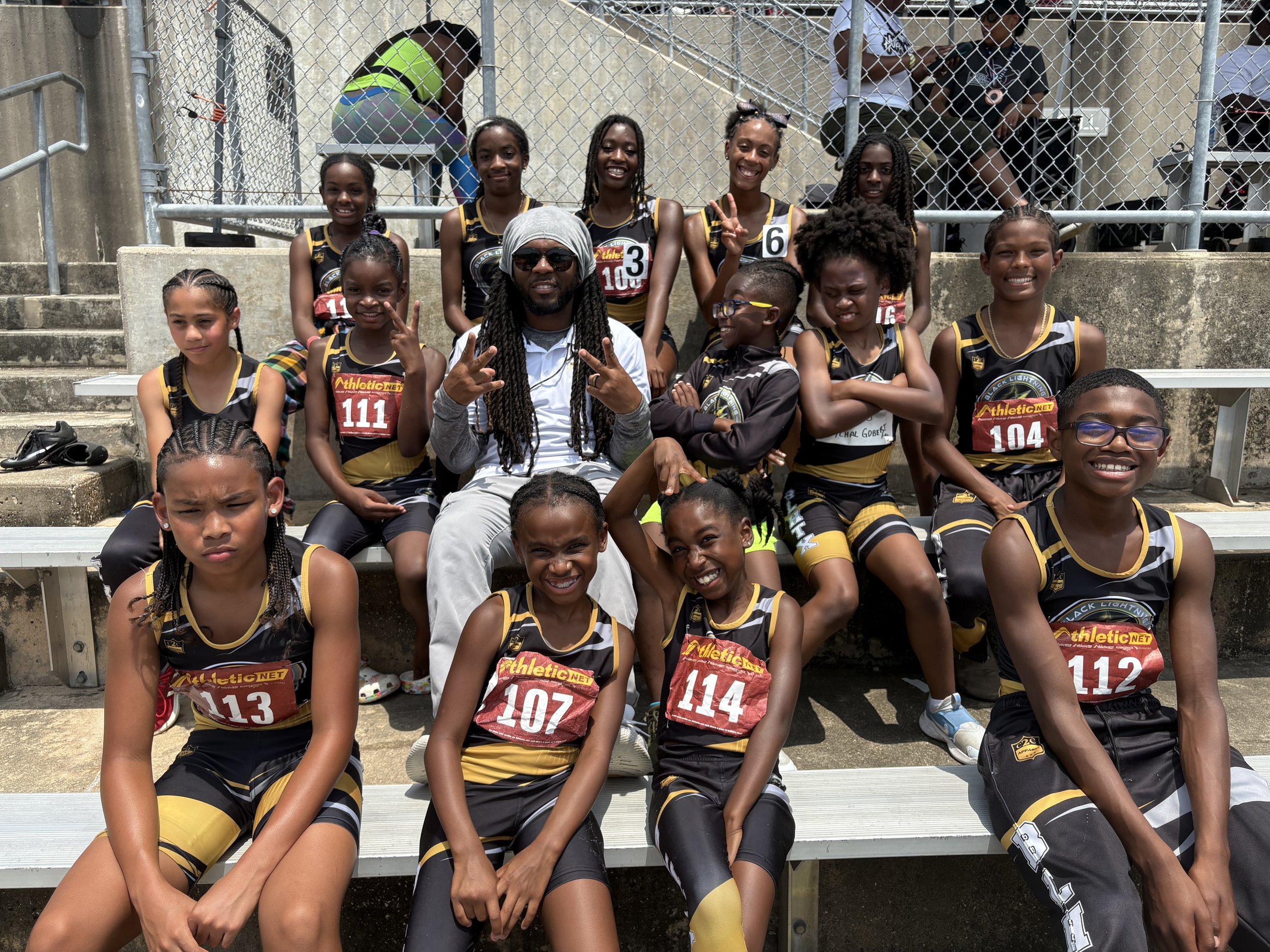 A group of young female track athletes in black and yellow uniforms sitting on bleachers outdoors, posing with their coach who is making hand gestures. The athletes have race numbers on their uniforms, and some are smiling or making playful poses.