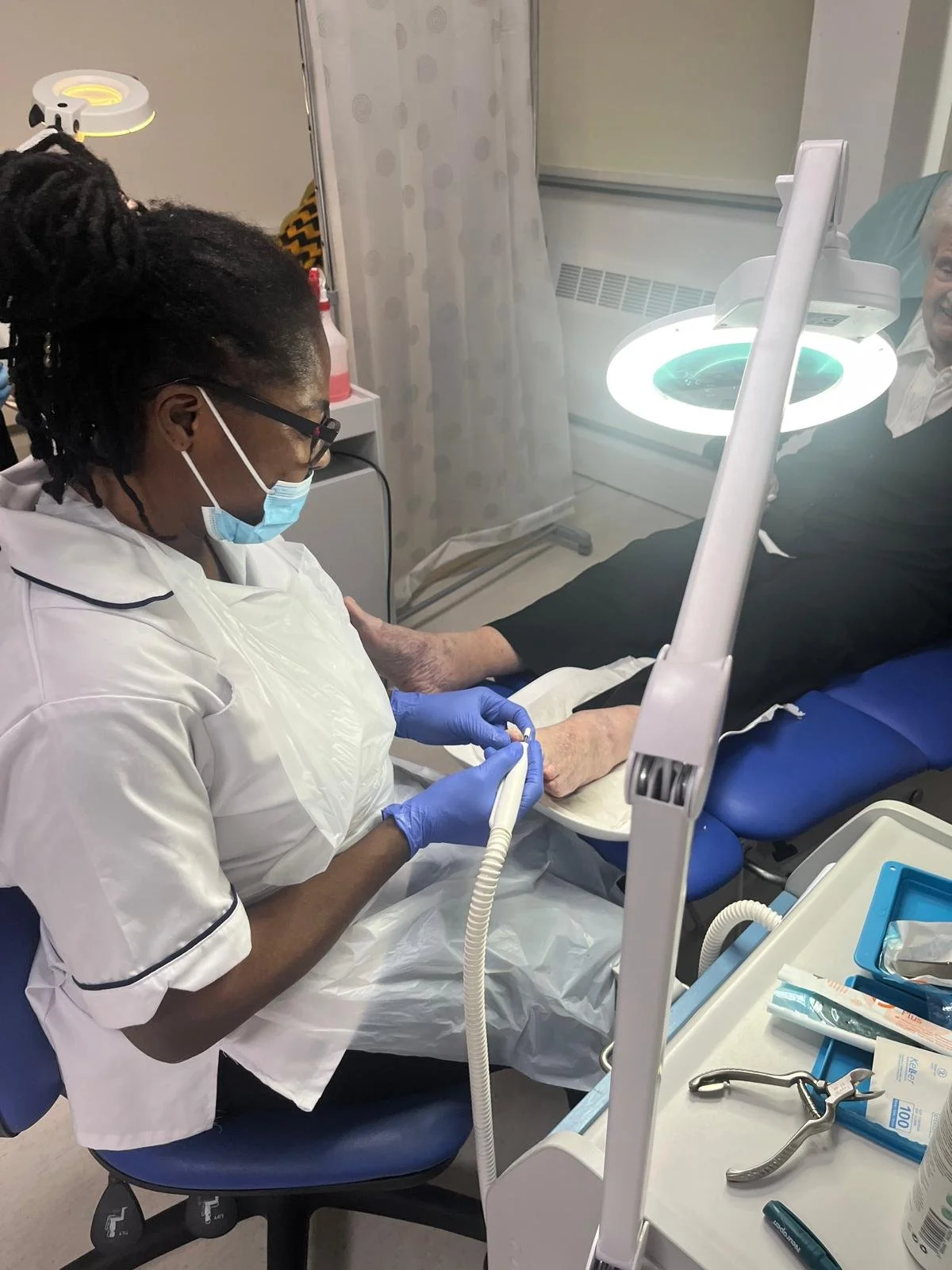 A nurse in a white uniform and face mask performing a medical procedure on an elderly man lying on a hospital bed, with medical tools and equipment nearby.