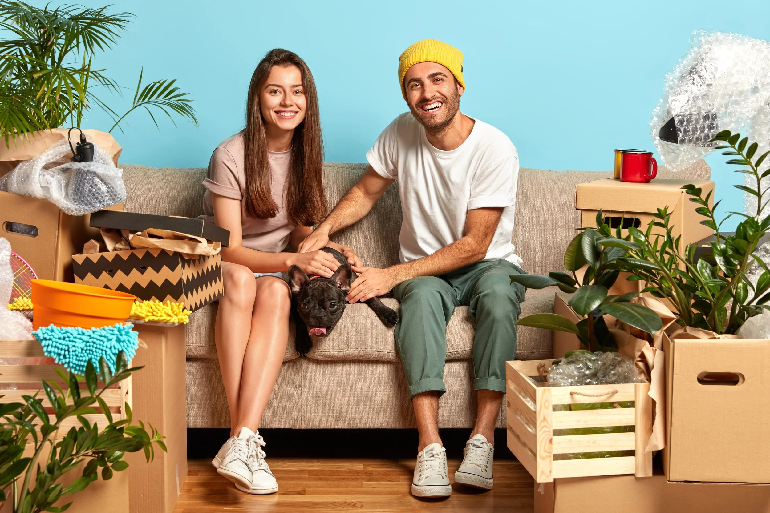 A young couple sitting on a sofa surrounded by moving boxes and plants, with a black French bulldog puppy, smiling and enjoying the moment.