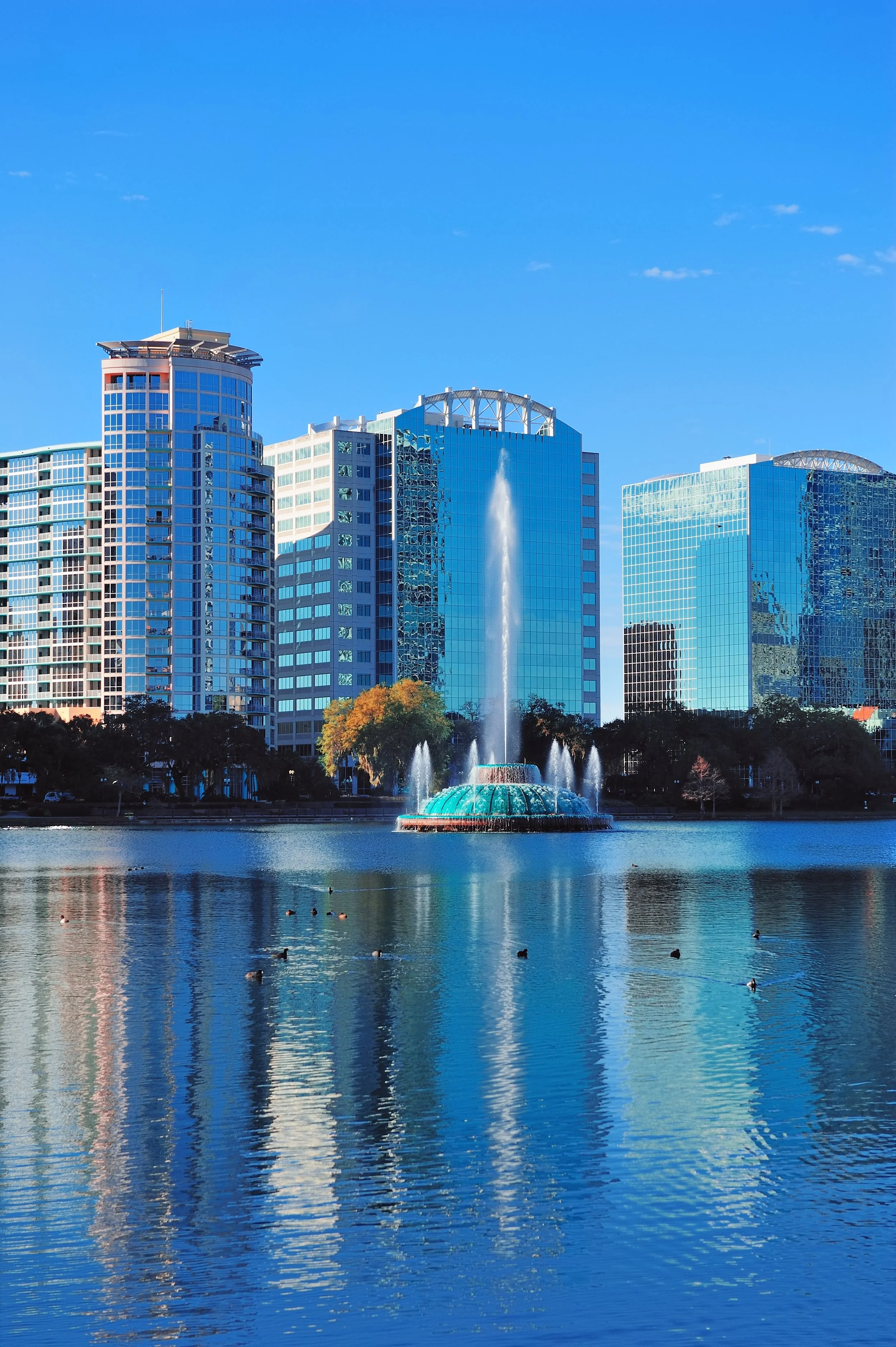 City skyline with modern glass buildings and a large fountain in a lake, under a clear blue sky.