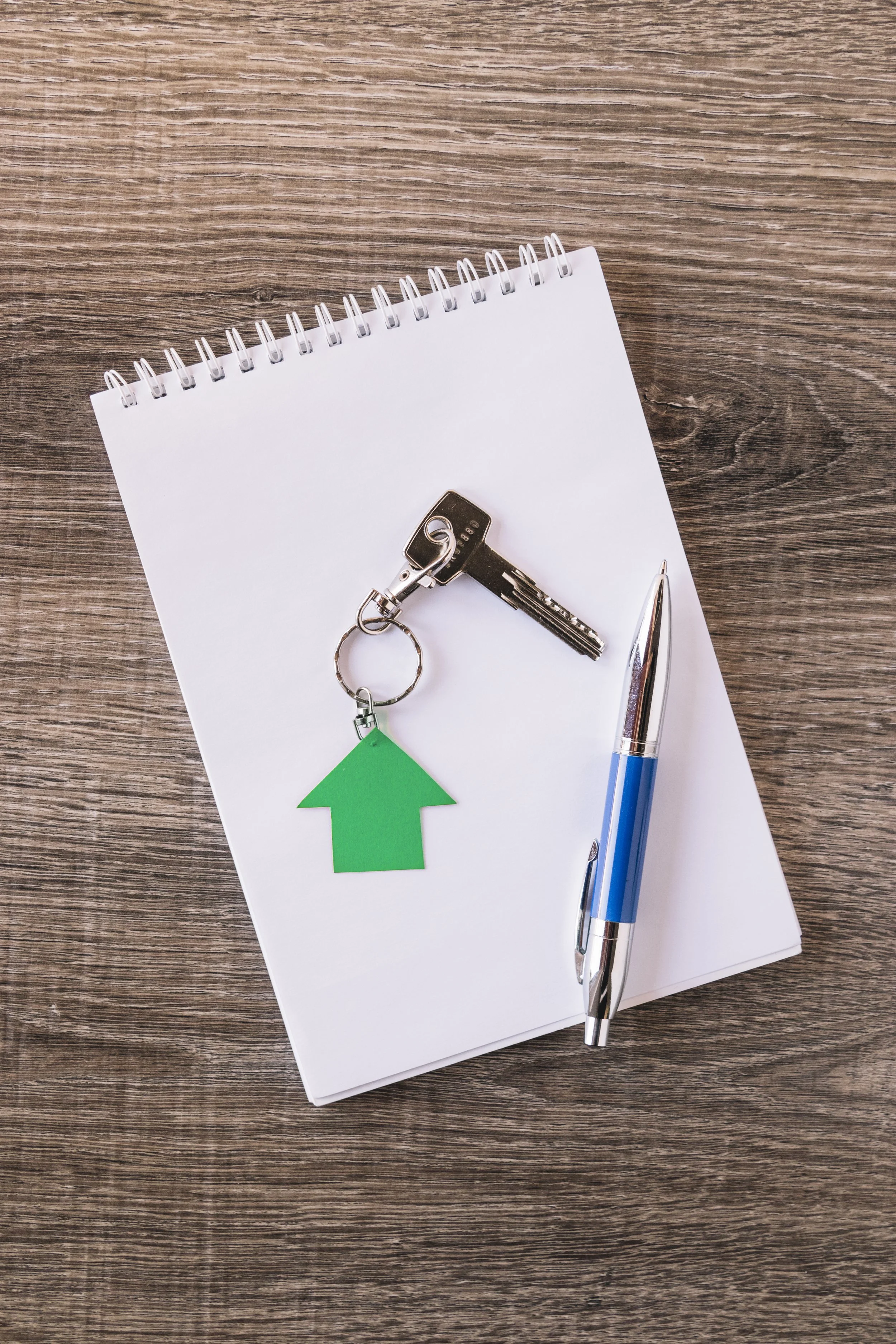 A set of keys with a green house-shaped keychain, a blue pen, and a blank notepad on a wooden surface.