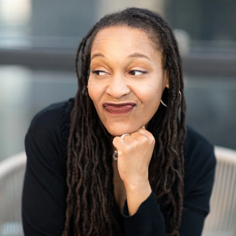 A woman with long dreadlocks and a nose ring, wearing a black top, sitting outdoors with her chin resting on her hand, looking to the side with a slight smile.