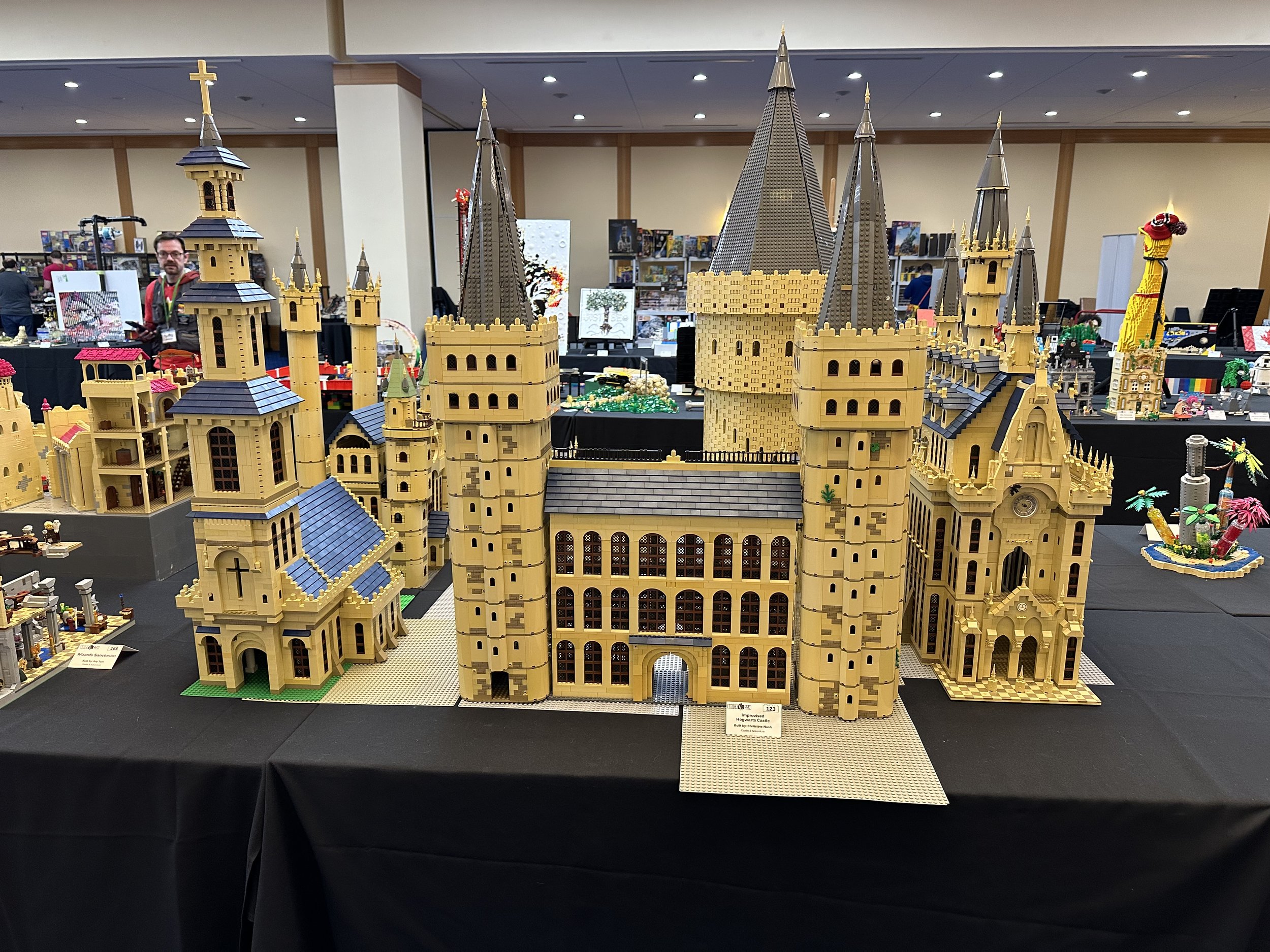 Detailed LEGO model of a castle with multiple towers, turrets, and architectural features, displayed on a black table at a LEGO exhibition.