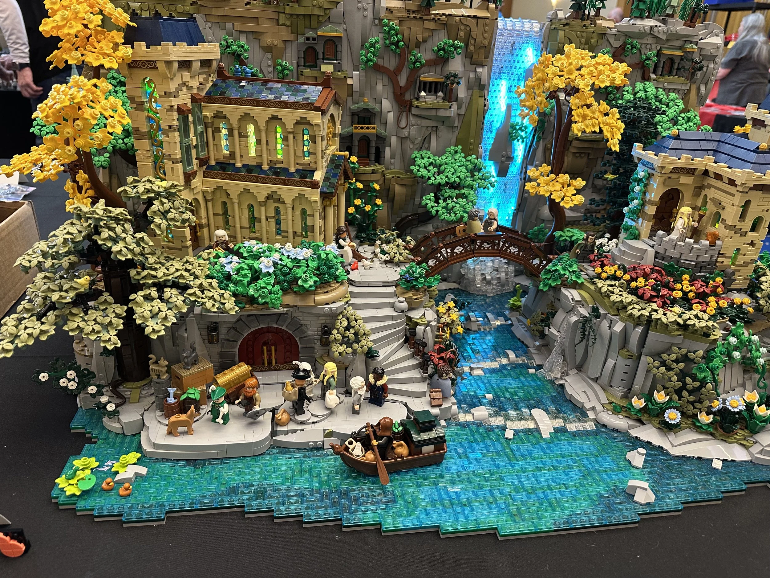 Lego display of a detailed fantasy landscape with waterfalls, a river, trees, flowers, and miniature figures including people and animals, depicting a vibrant scene with buildings and bridges.