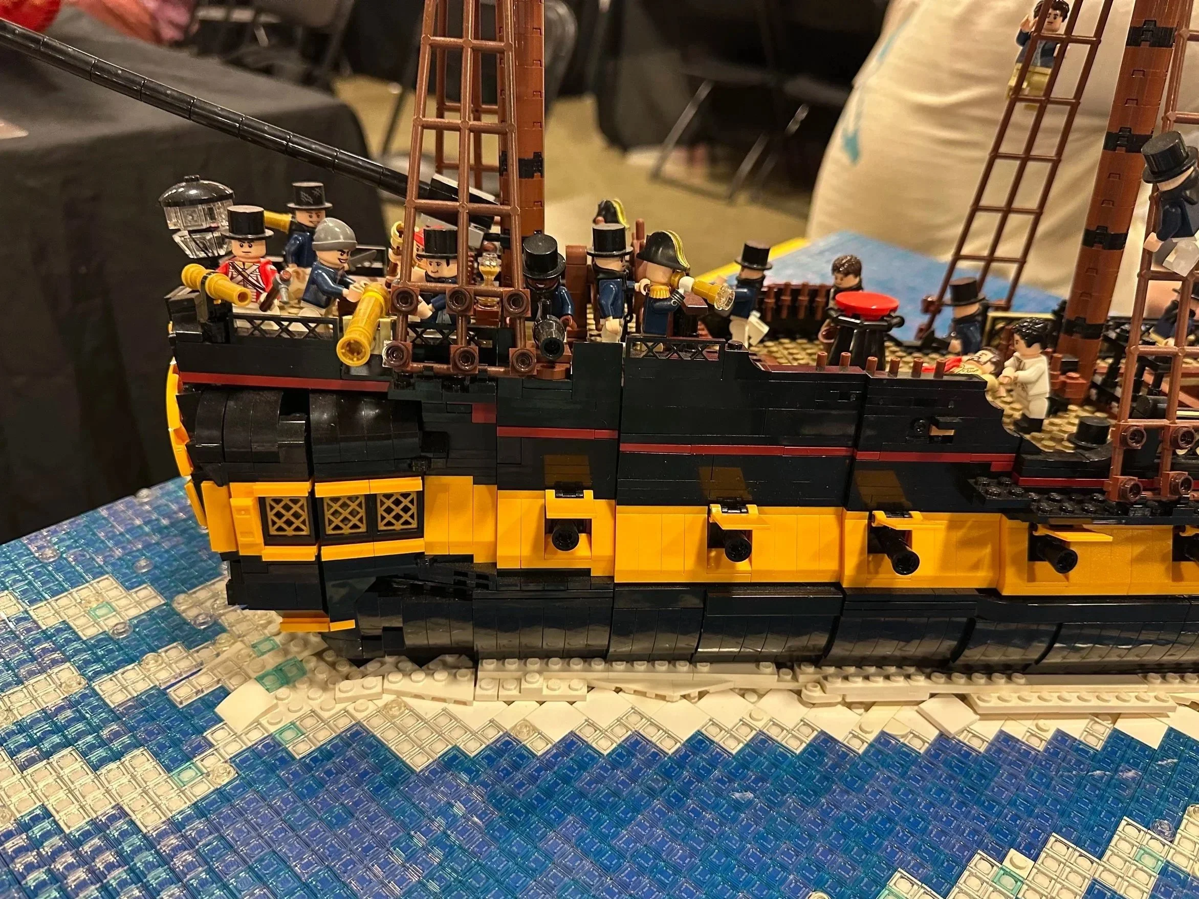 LEGO model of a historic sailing ship with miniature LEGO figures onboard, displayed on a table with a blue woven covering.