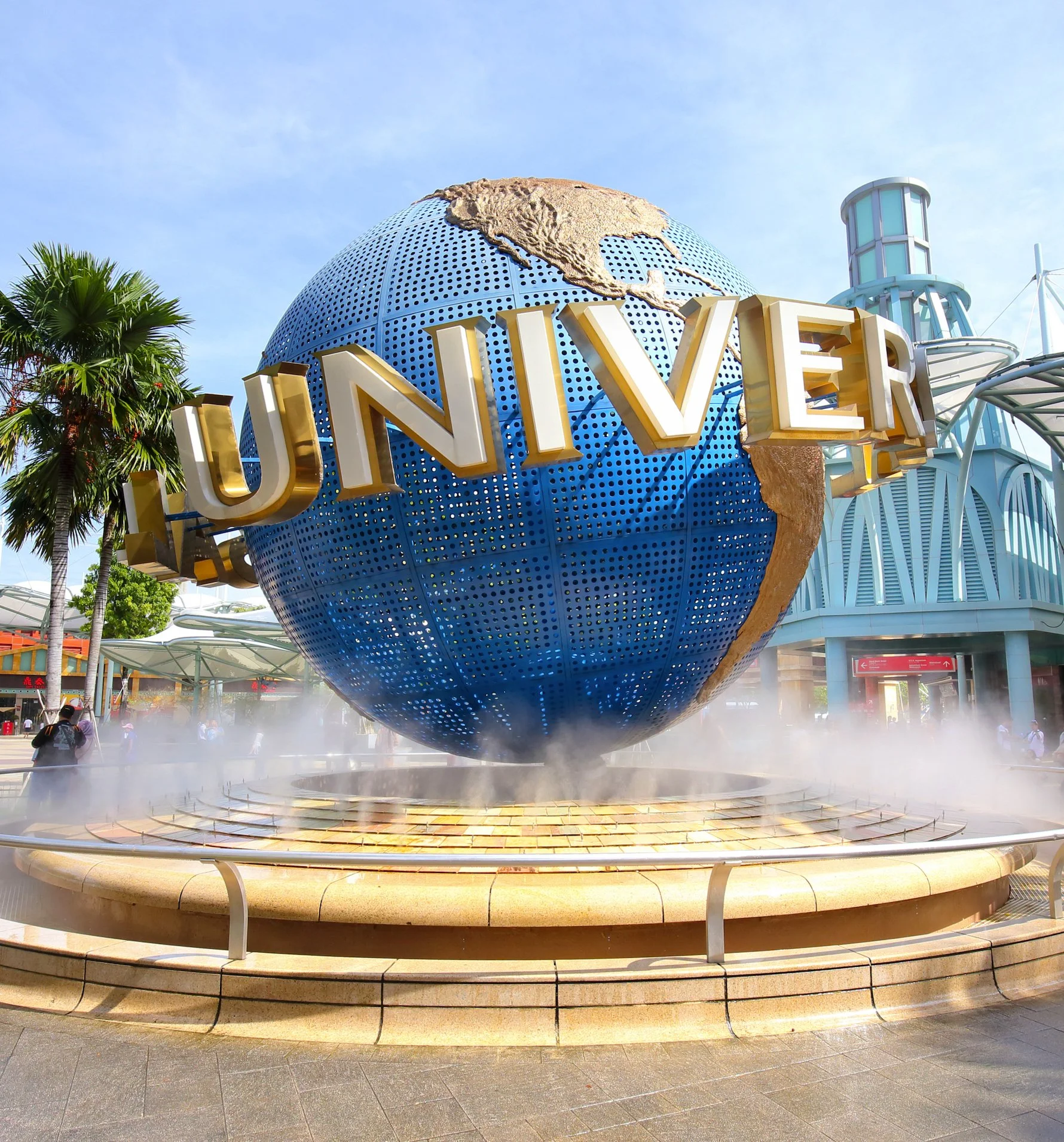 Universal Studios globe fountain with the word 'UNIVERSAL' across the globe, located at Universal Studios theme park, with palm trees and park structures in the background.