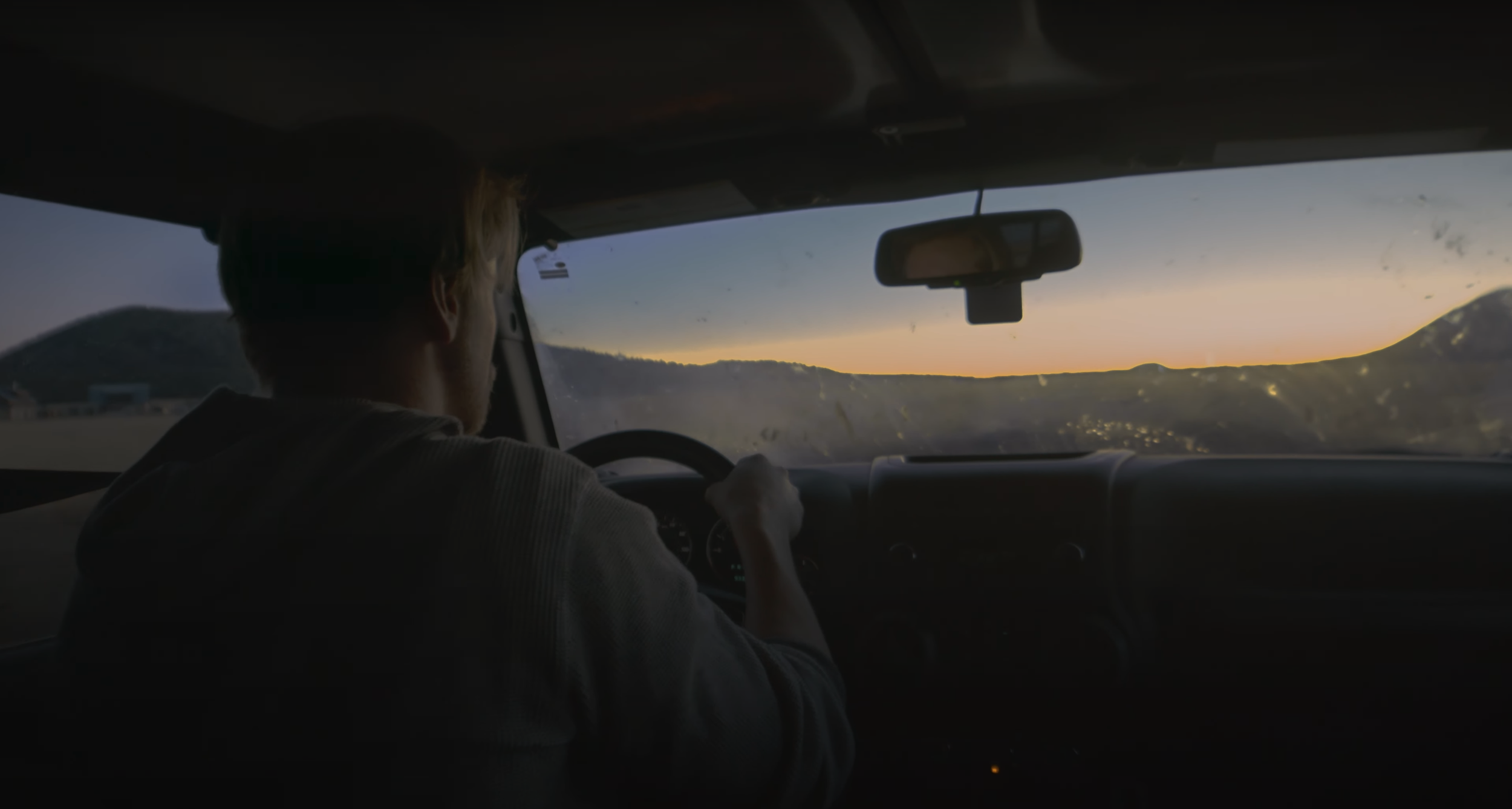 The view of a man driving from the back seat. The sun can be seen rising through the windshield.