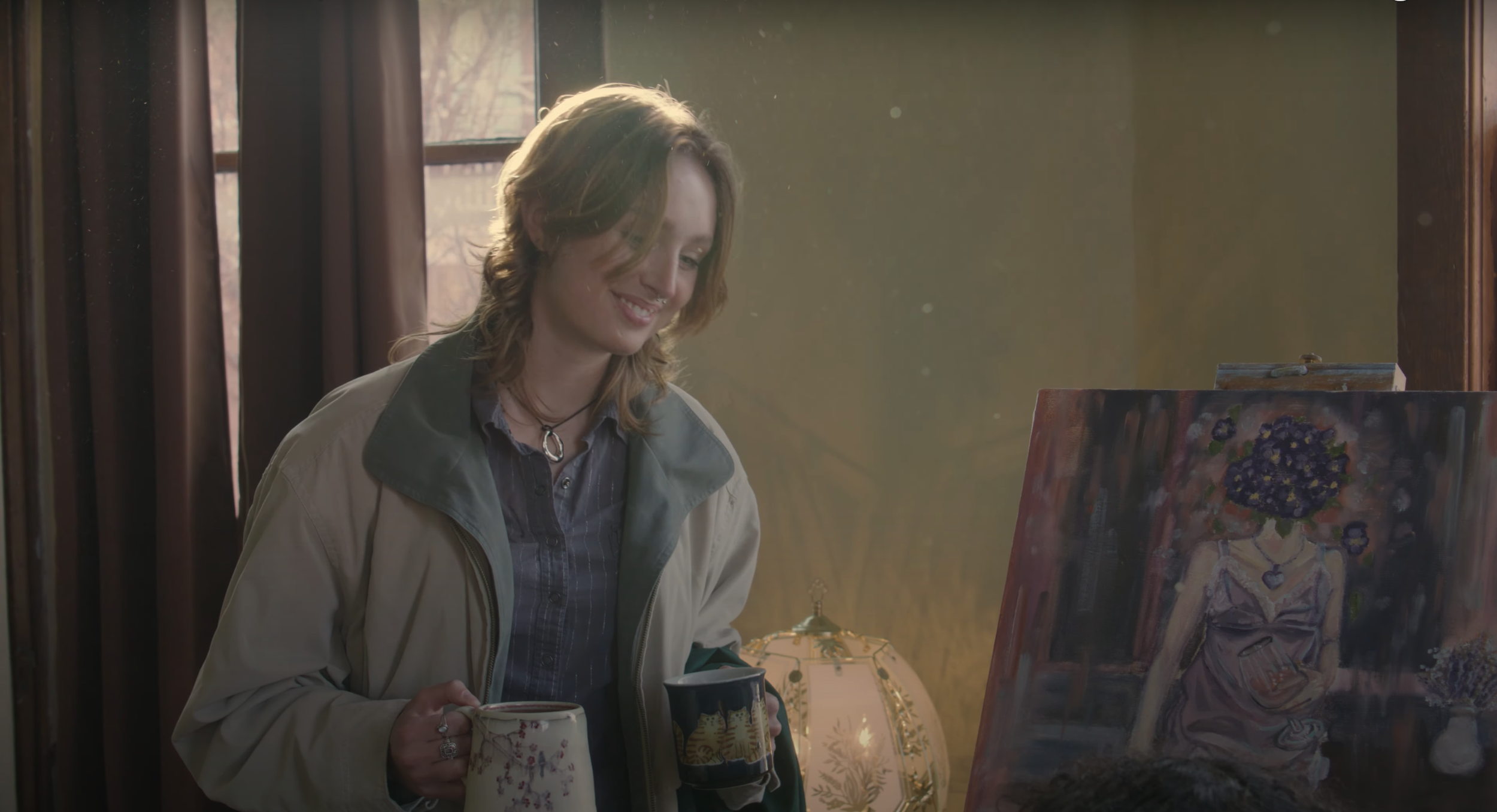 A woman holding two mugs smiles. Her hair catches the sunlight.