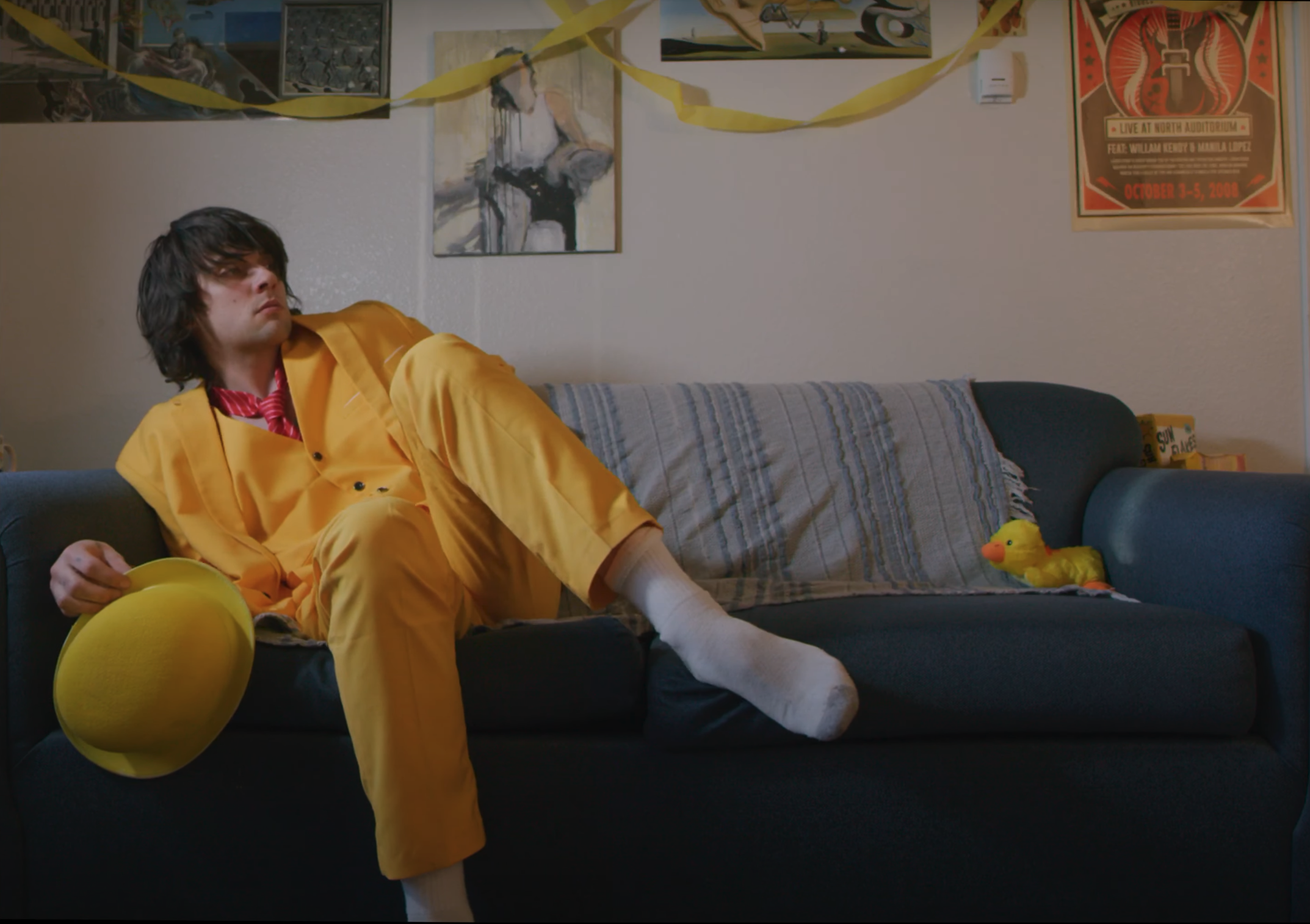 A man with dark, unkempt hair sits awkwardly on a couch. He is wearing a red tie and an all-yellow suit, and holding a matching yellow bowler hat.