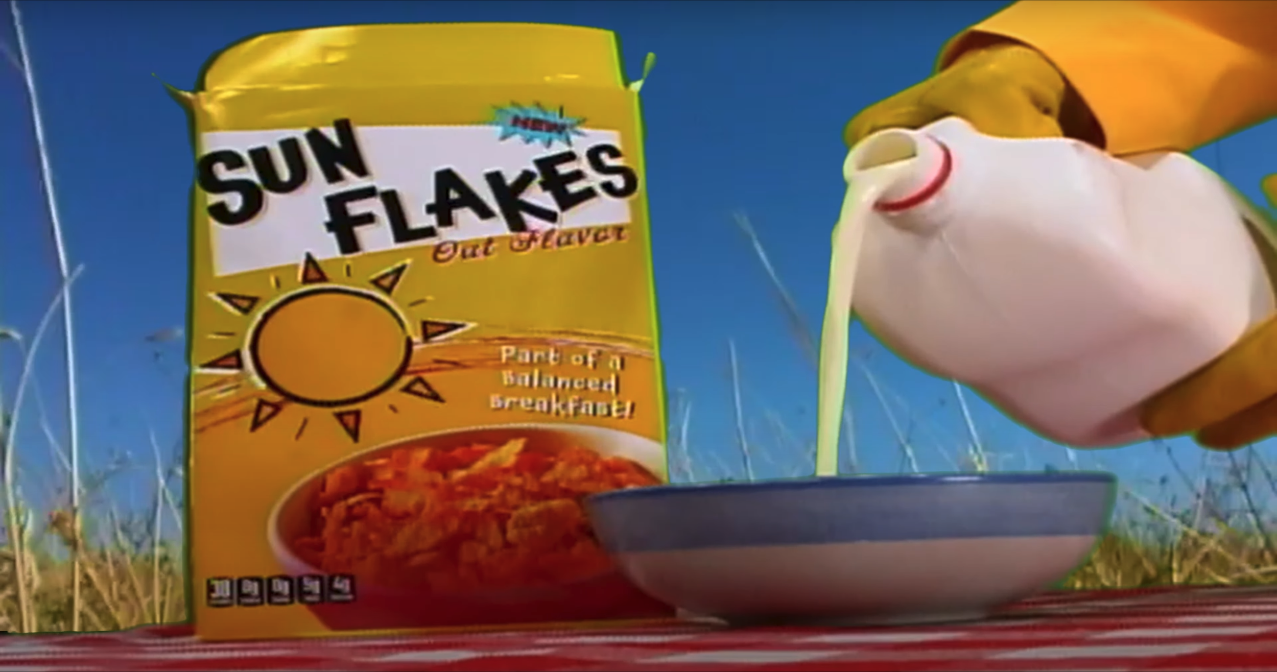On a red and white gingham table cloth sits a yellow cereal box labeled "Sun Flakes", a blue cereal bowl, and a yellow-gloved hand pourig a gallon of milk. In the background, a yellow wheat field stands out against the bright blue sky.
