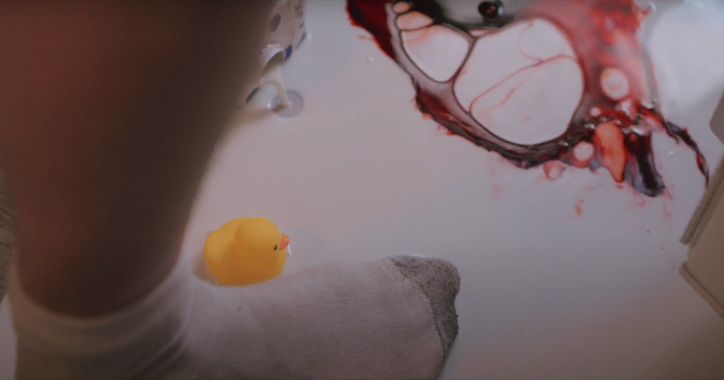 Milk and blood pour out onto the floor and is illuminated by the light from a refrigerator. In the puddle is a yellow rubbery ducky and a foot wearing a crew sock.