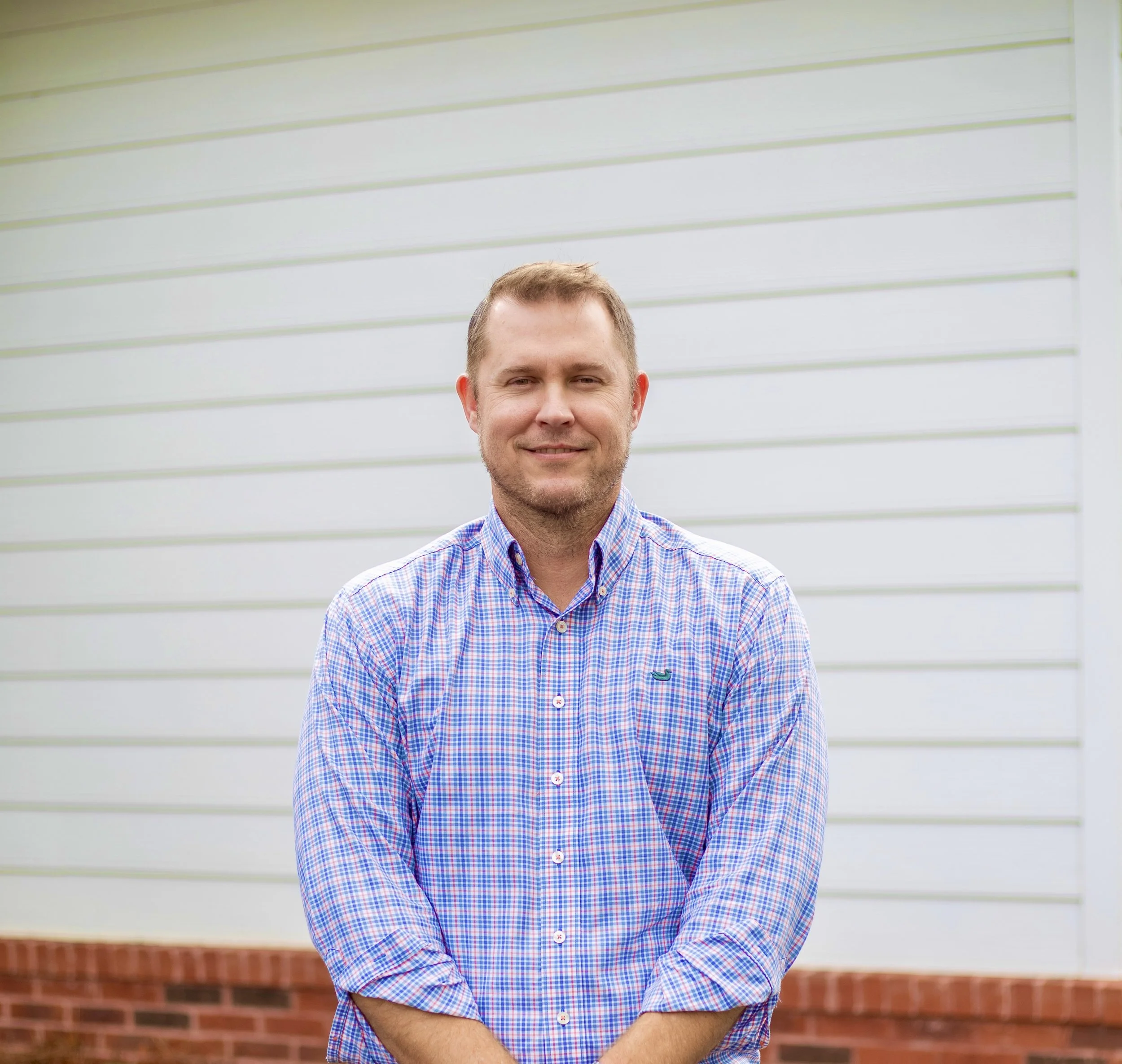 A man wearing a colorful checkered shirt standing outdoors in front of a white wall with horizontal siding.