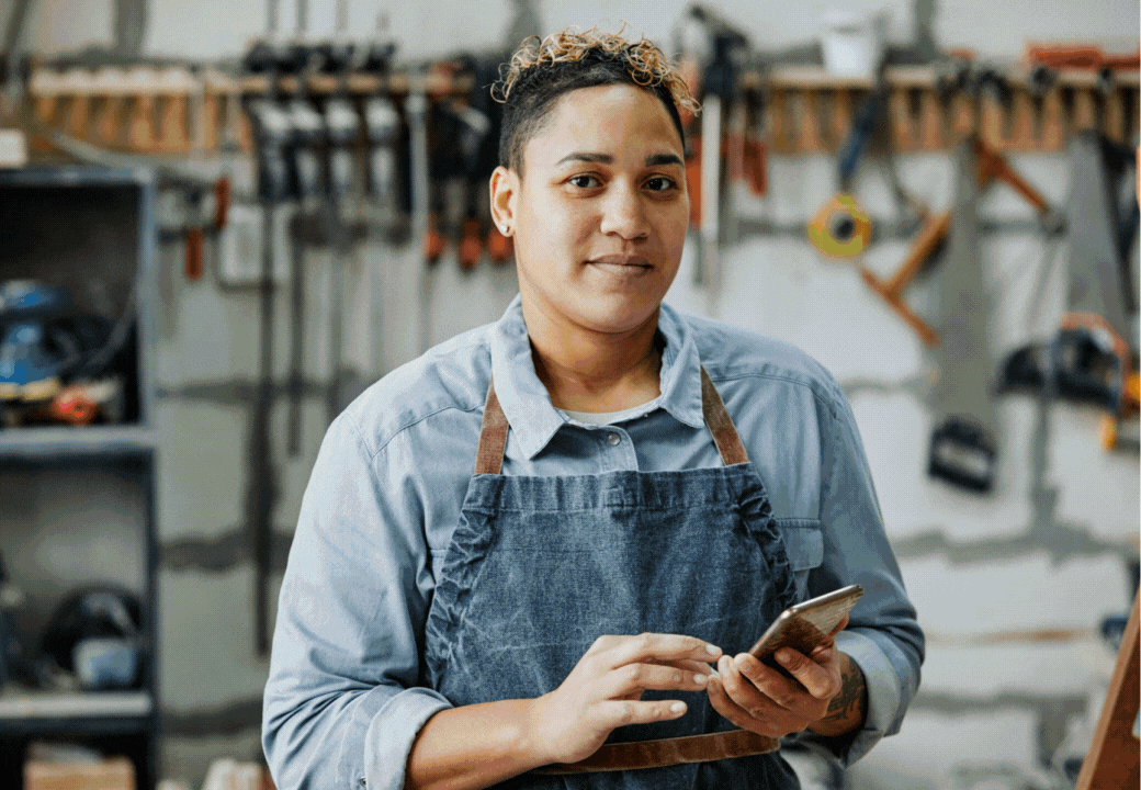 A woman in a workshop holds a smartphone, wearing a denim apron and a light blue shirt, with tools hanging on the wall behind her.