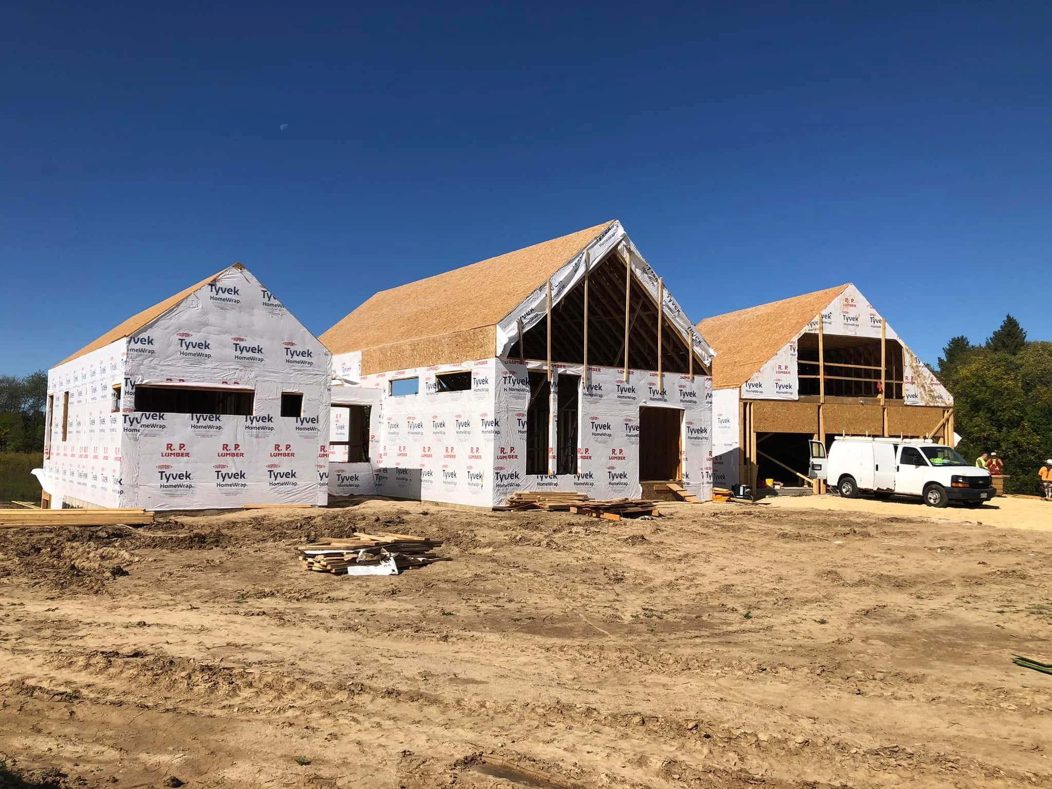 Under construction house with partially installed roof and exterior walls wrapped in Tyvek HomeWrap, with a white van parked in front and construction workers on site.