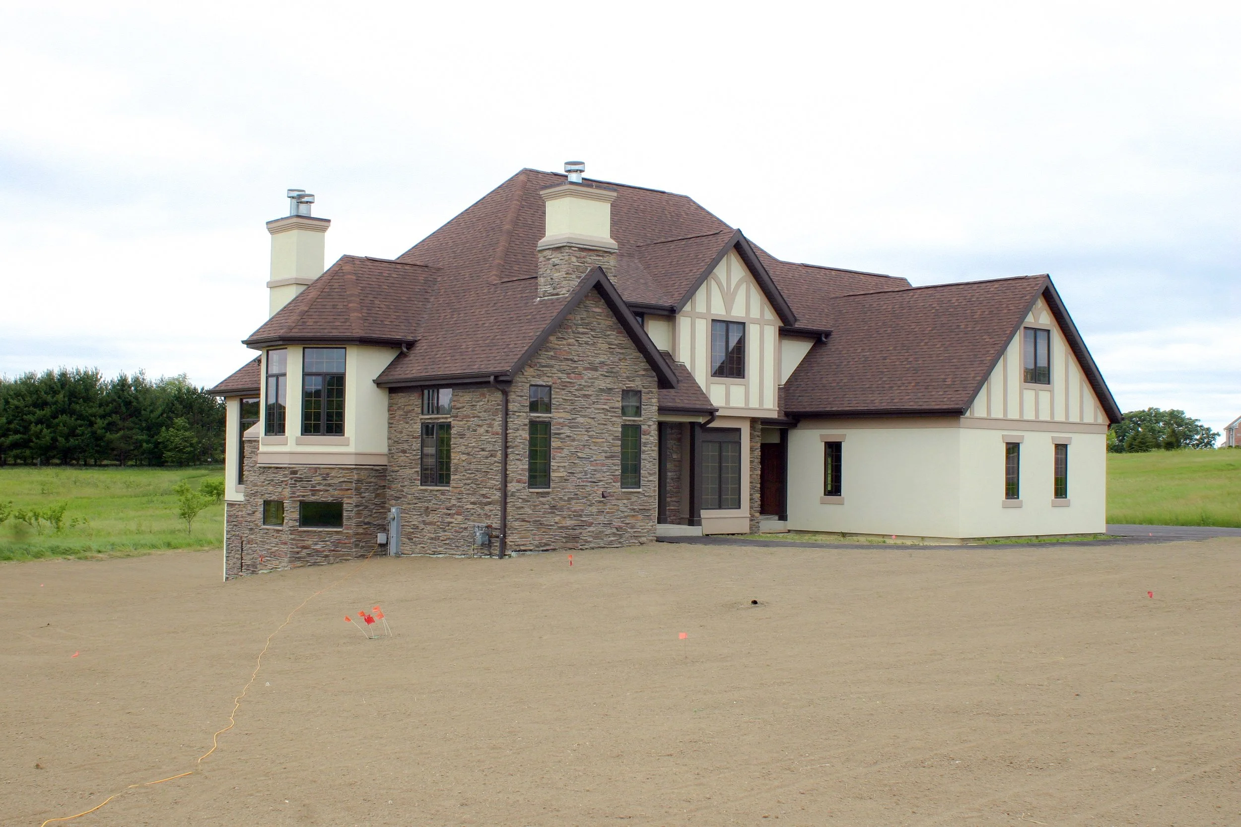 A newly constructed house with a mix of stone and siding exterior, large windows, and a multi-gabled roof, situated on a large patch of bare dirt, with green fields and trees in the background.
