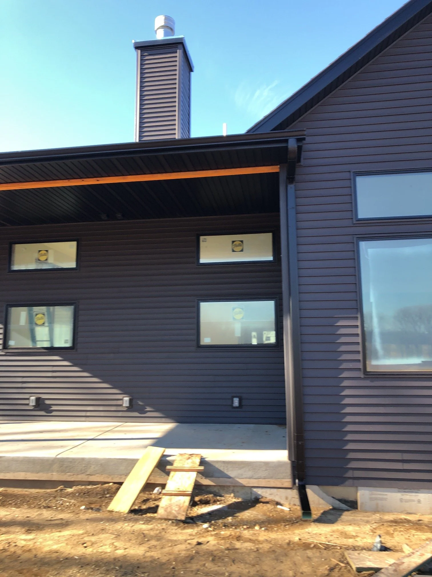 Exterior of a modern house under construction, featuring dark siding, a chimney, multiple windows, and a concrete foundation with construction materials on the ground.