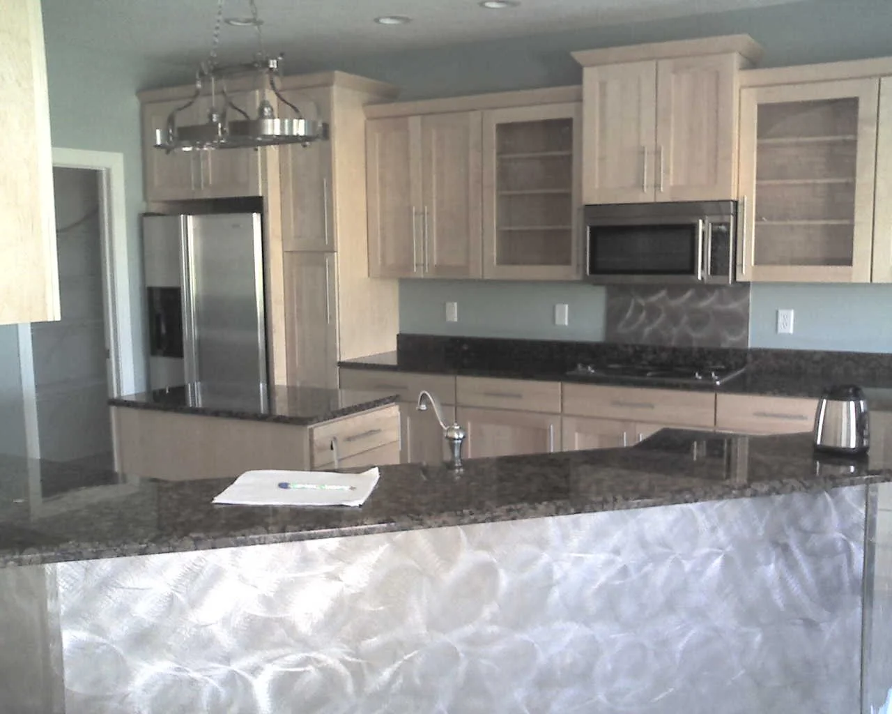 Modern kitchen with light wood cabinets, stainless steel appliances, black granite countertops, and a decorative backsplash.
