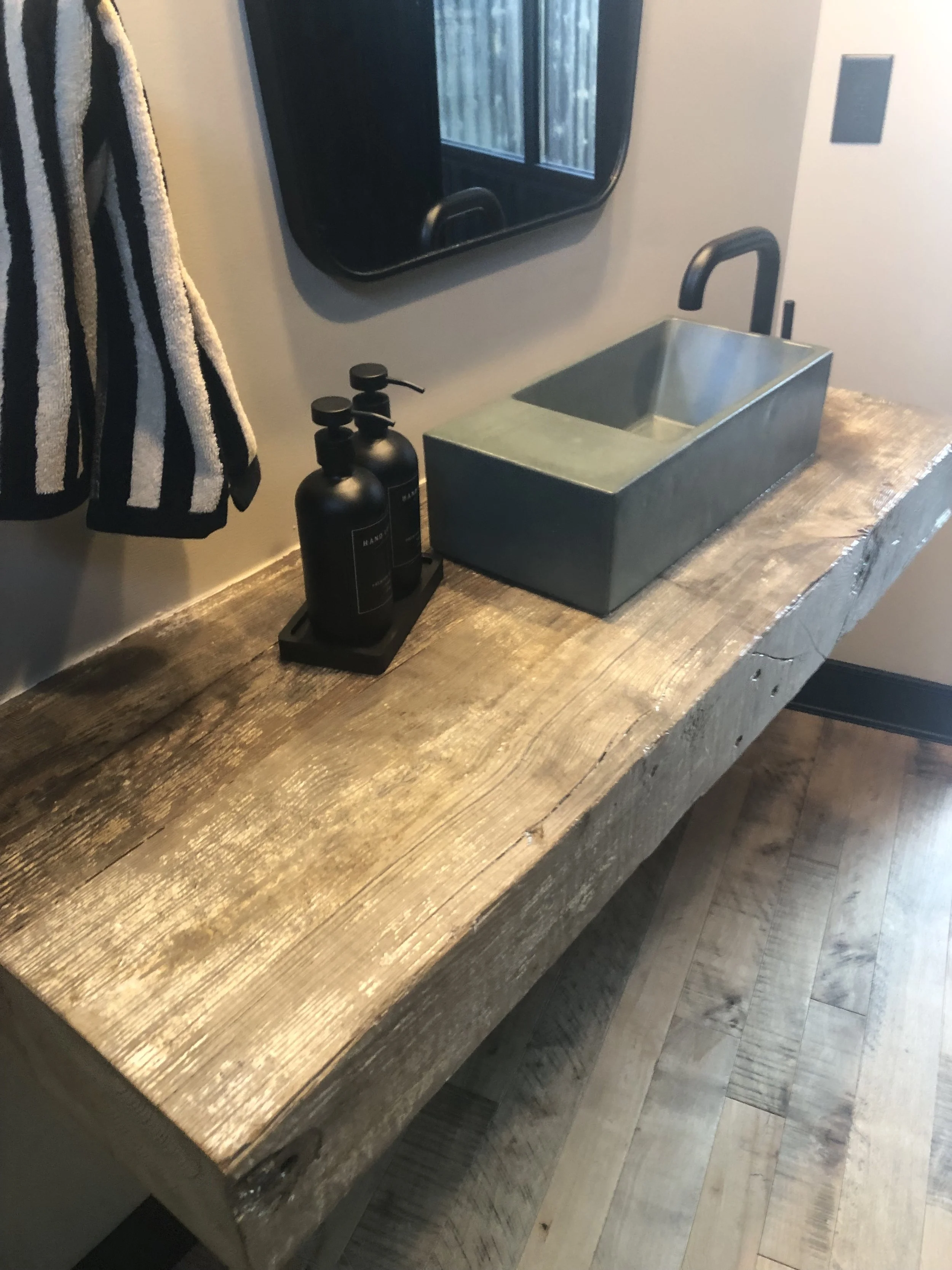 Rustic wooden bathroom vanity with a rectangular stainless steel sink, black faucet, and black soap dispensers, with a window, black framed mirror, and striped towel hanging nearby.